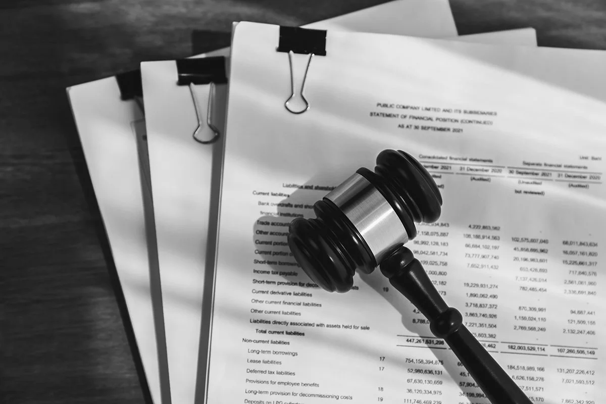 A gavel resting on financial documents with charts and data, held down by black binder clips on a wooden table.