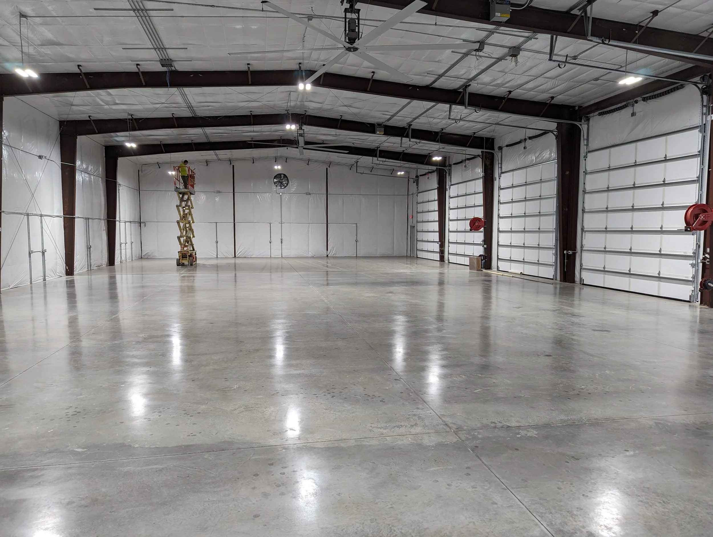 An empty industrial warehouse with polished concrete floors, white insulated walls, and high ceilings with exposed beams. There is a person on a scissor lift working near the ceiling fans and ceiling lights. Large garage doors are visible on the righ