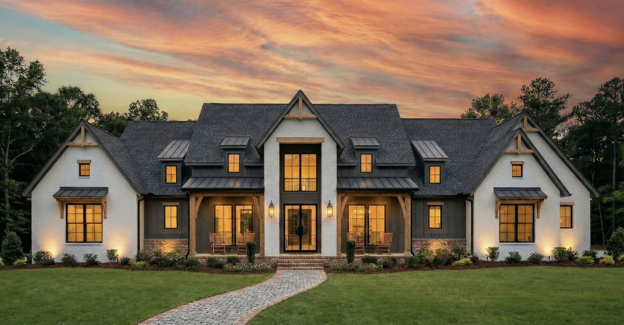 Large modern house with a gabled roof, illuminated windows, surrounded by a green lawn and trees, during sunset.