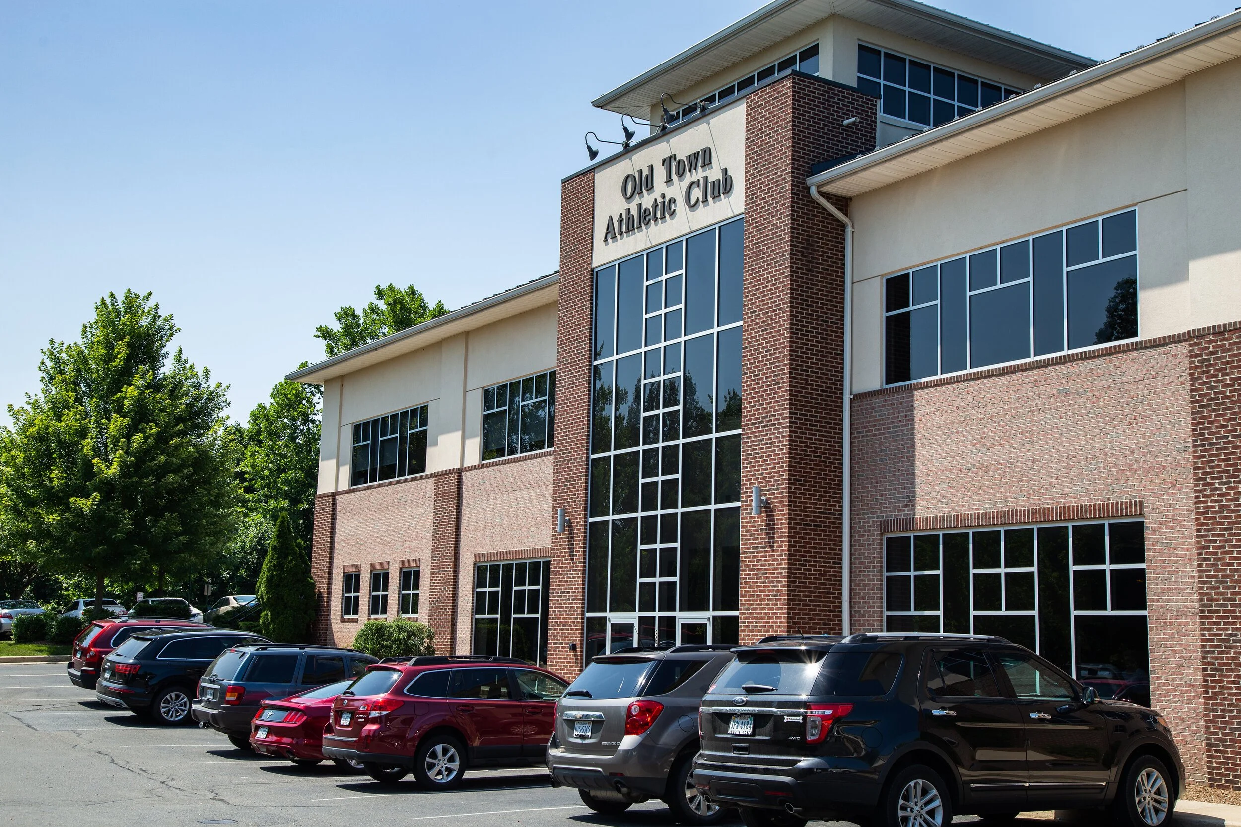Front view of the Old Town Athletic Club building with parking lot and several cars parked in front, trees on the left side, and a clear blue sky.