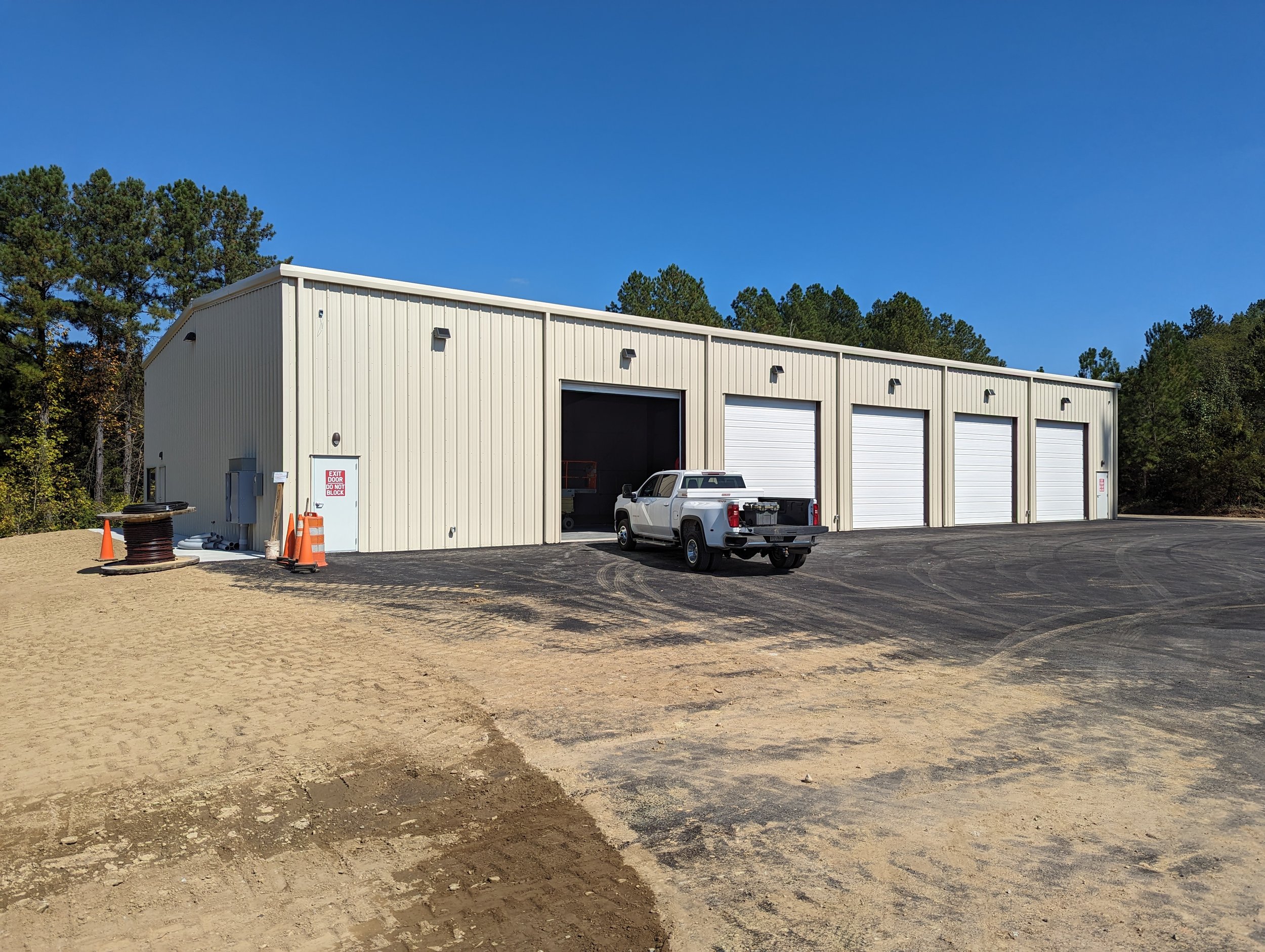 A large beige metal warehouse with four garage doors, a pickup truck parked outside, construction cones, and a dirt area in front, surrounded by trees under a clear blue sky.