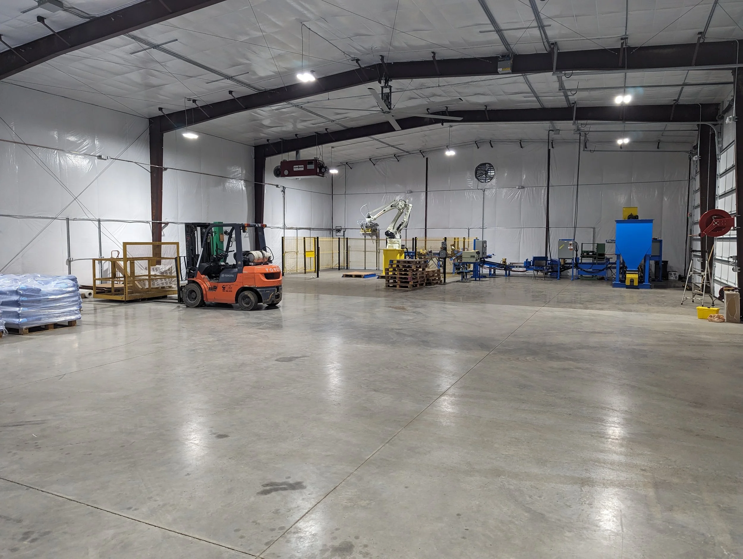 Interior of a warehouse or industrial space with equipment, pallets, and a forklift, with a high ceiling, lighting, and minimal decoration.