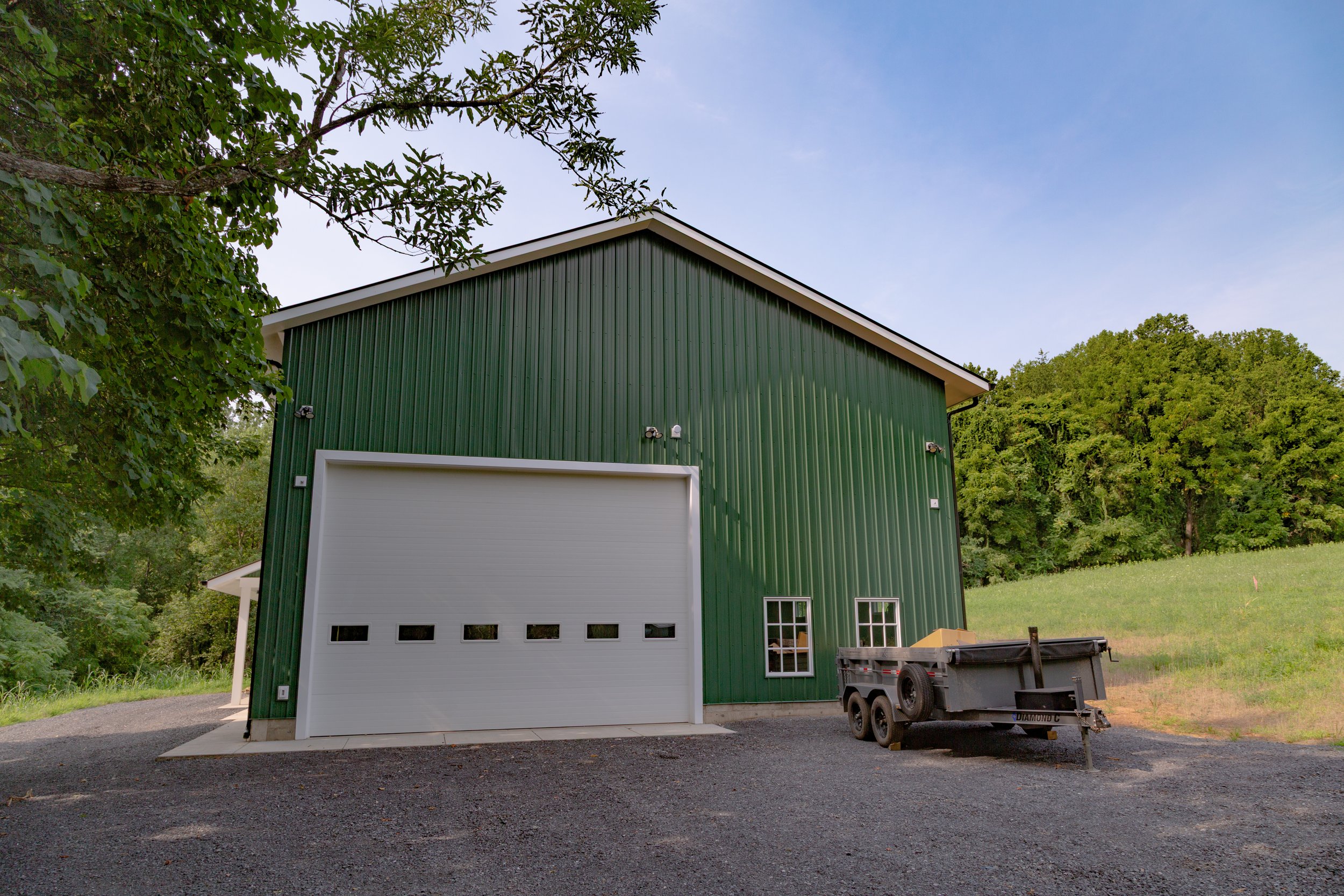 Green metal building with a large white garage door, surrounded by trees and a gravel driveway, with a trailer parked in front.
