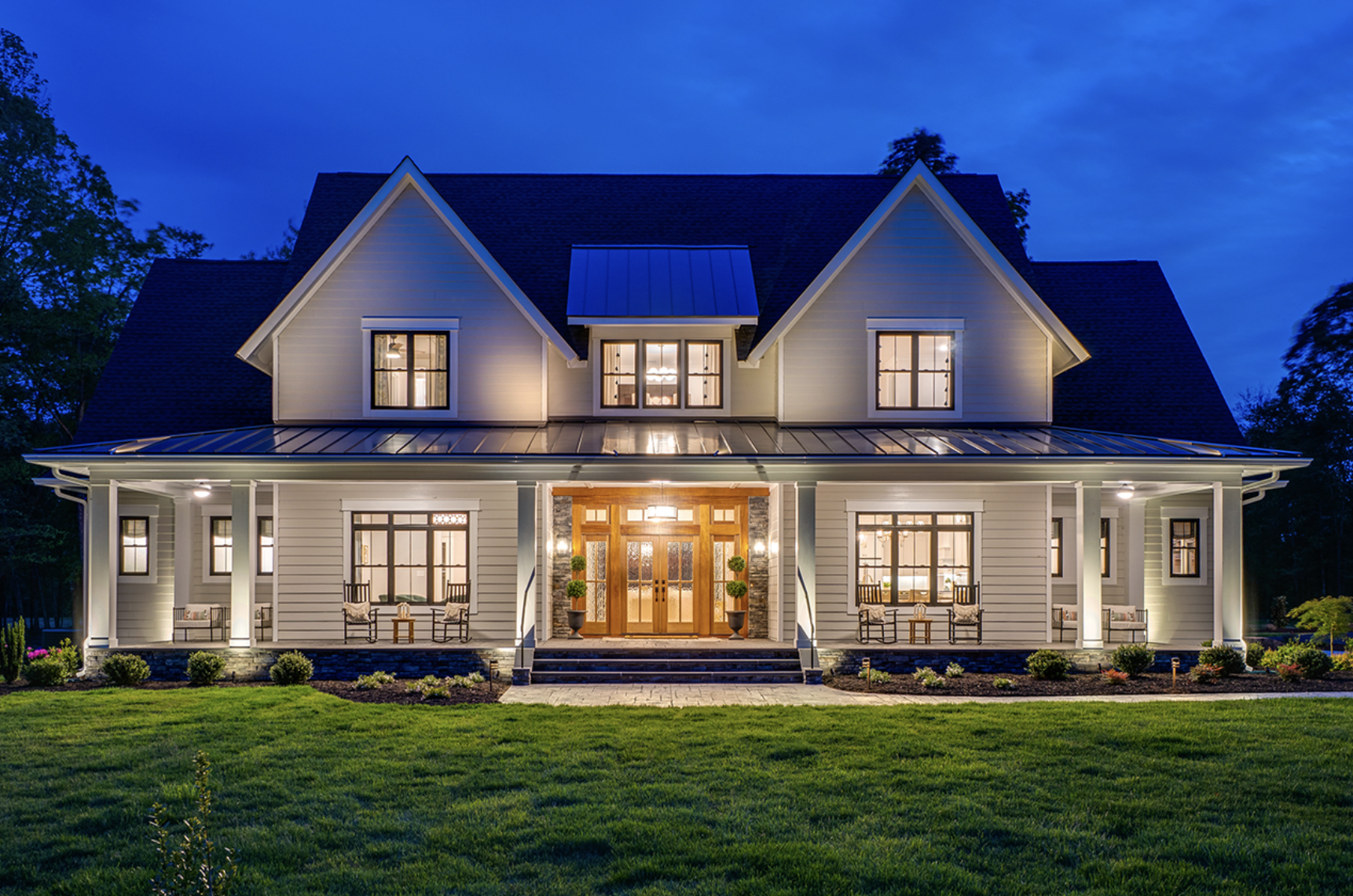 A large, two-story suburban house illuminated at night, featuring a front porch with seating, multiple windows with warm lighting, and a well-manicured lawn.