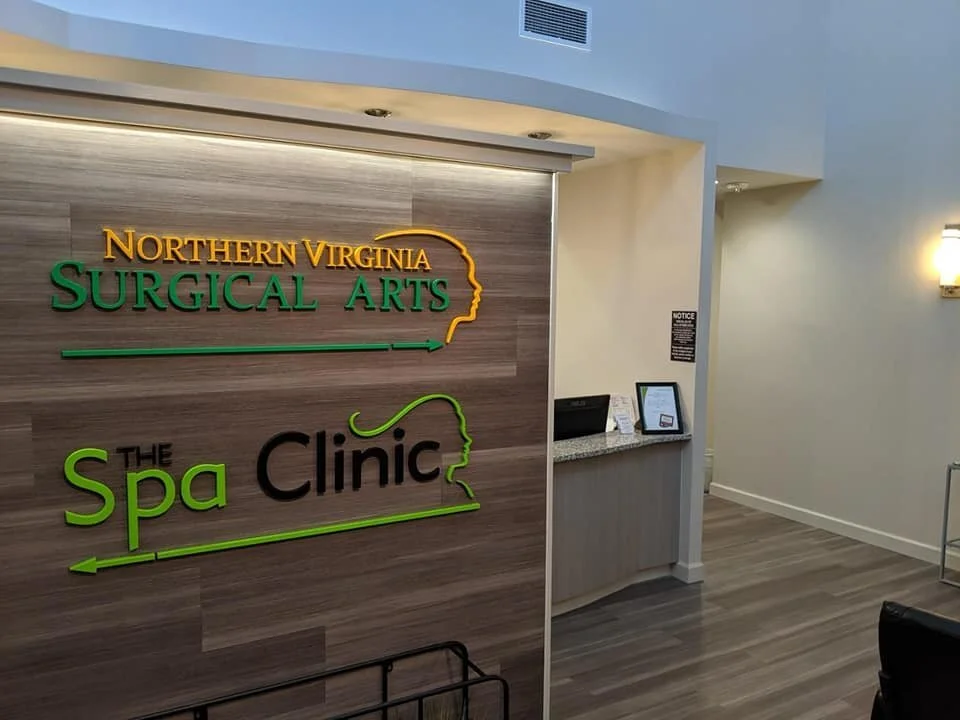 Interior of a medical or spa clinic with a wooden accent wall displaying the sign 'Northern Virginia Surgical Arts' and 'The Spa Clinic' in colorful letters, a small counter with a framed photo and papers, and a wall-mounted light fixture.