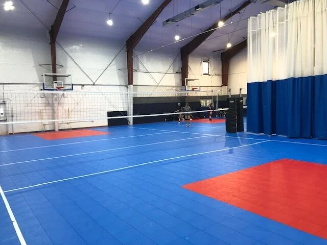 Indoor volleyball court with blue floor, red attack zone, white sideline, and two people playing volleyball near the net.