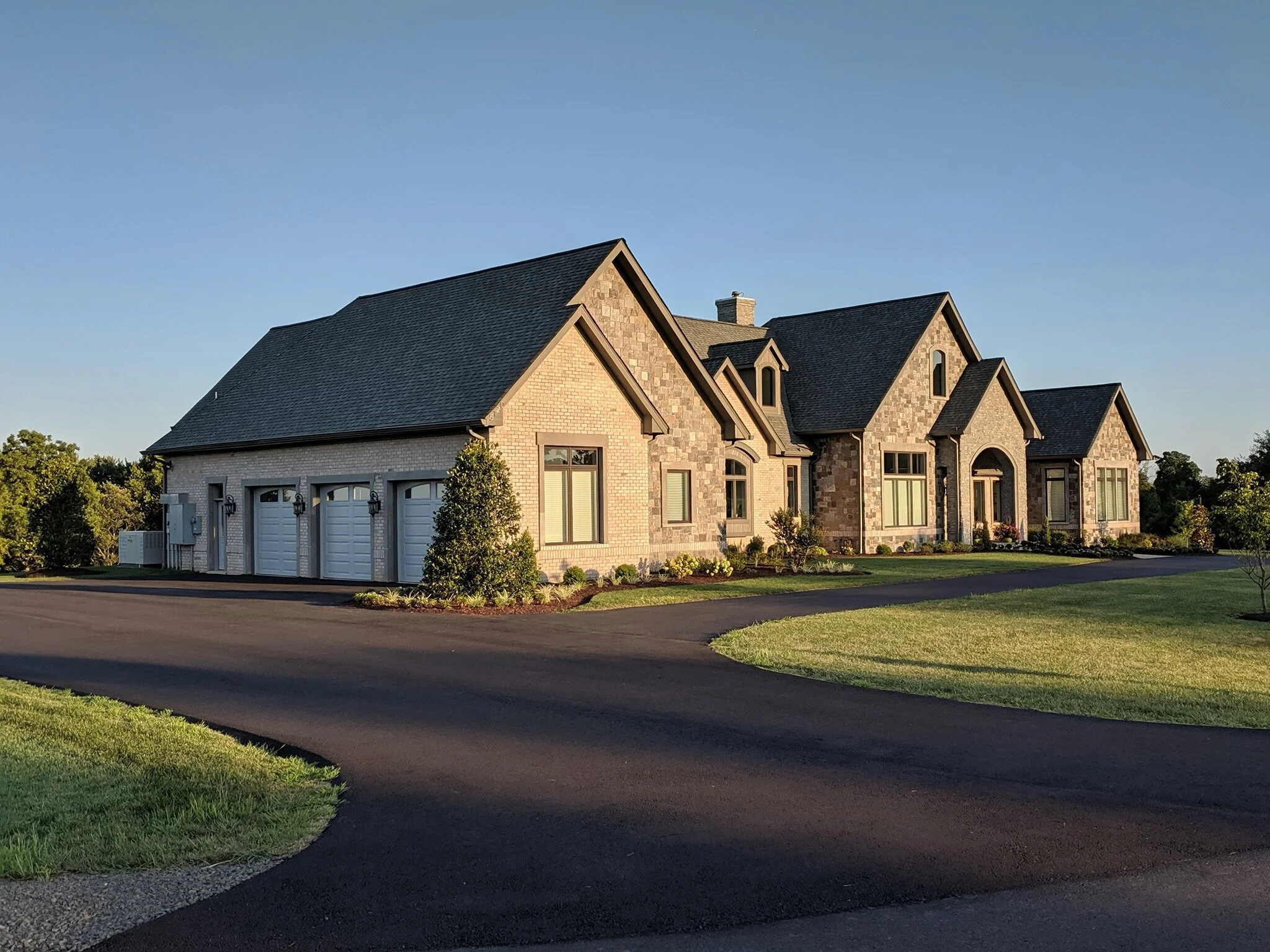 A large modern house with a gabled roof, brick and stone exterior, and multiple windows, surrounded by a well-maintained lawn and driveway under a clear blue sky.
