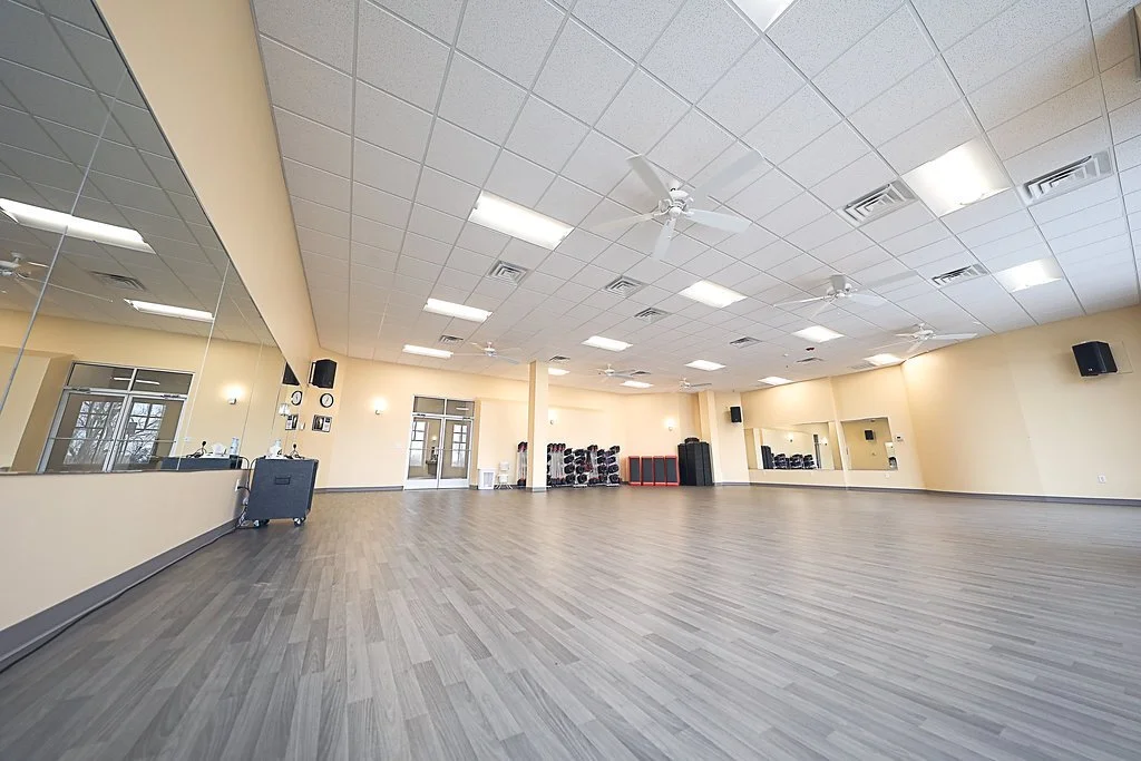 Empty fitness studio with wood flooring, large mirrors, ceiling fans, and exercise equipment stored against the back wall.