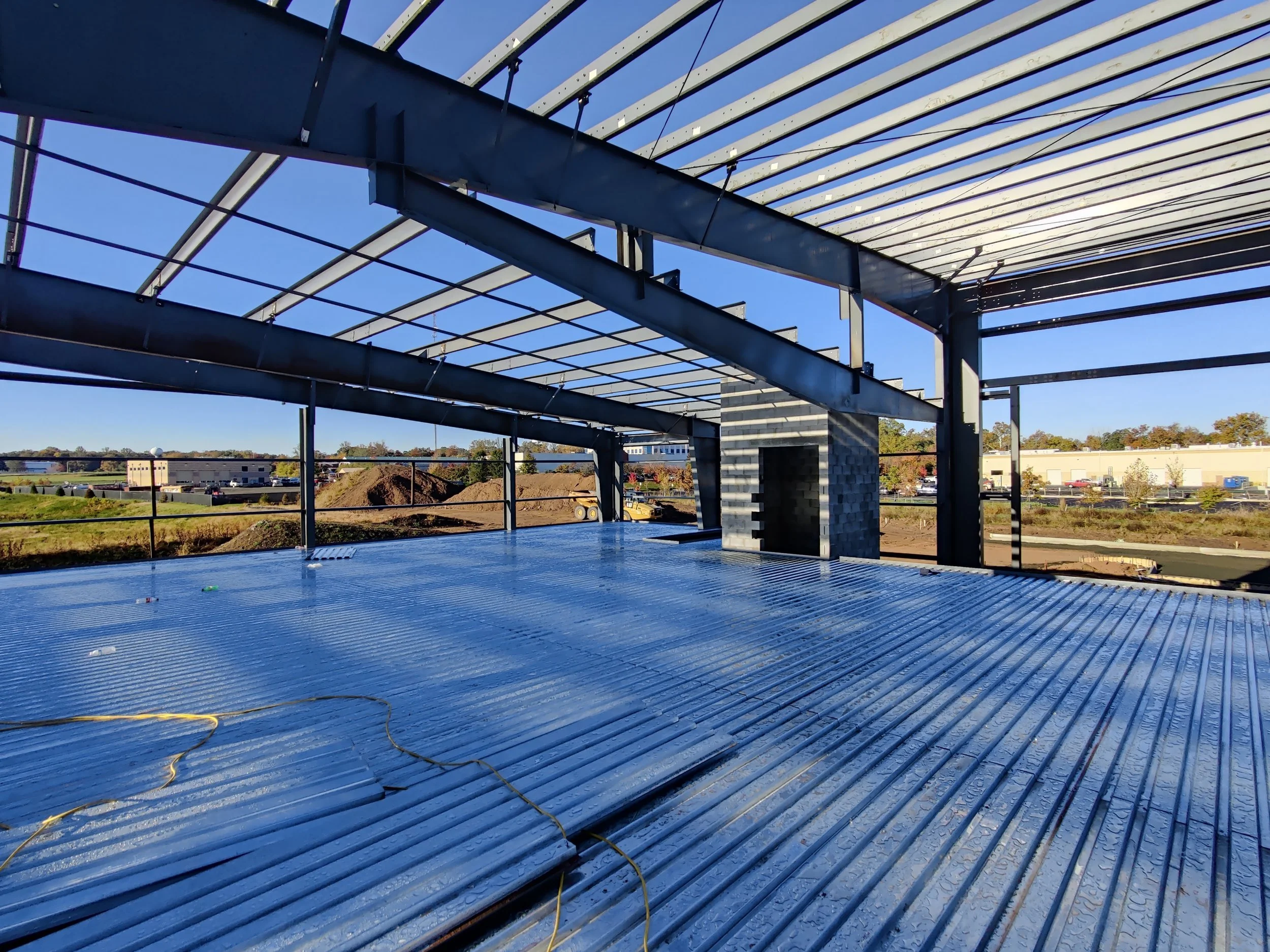 Under construction steel framework with a corrugated metal floor, view of a building under construction with a brick chimney, and a parking lot with cars and trees in the background.
