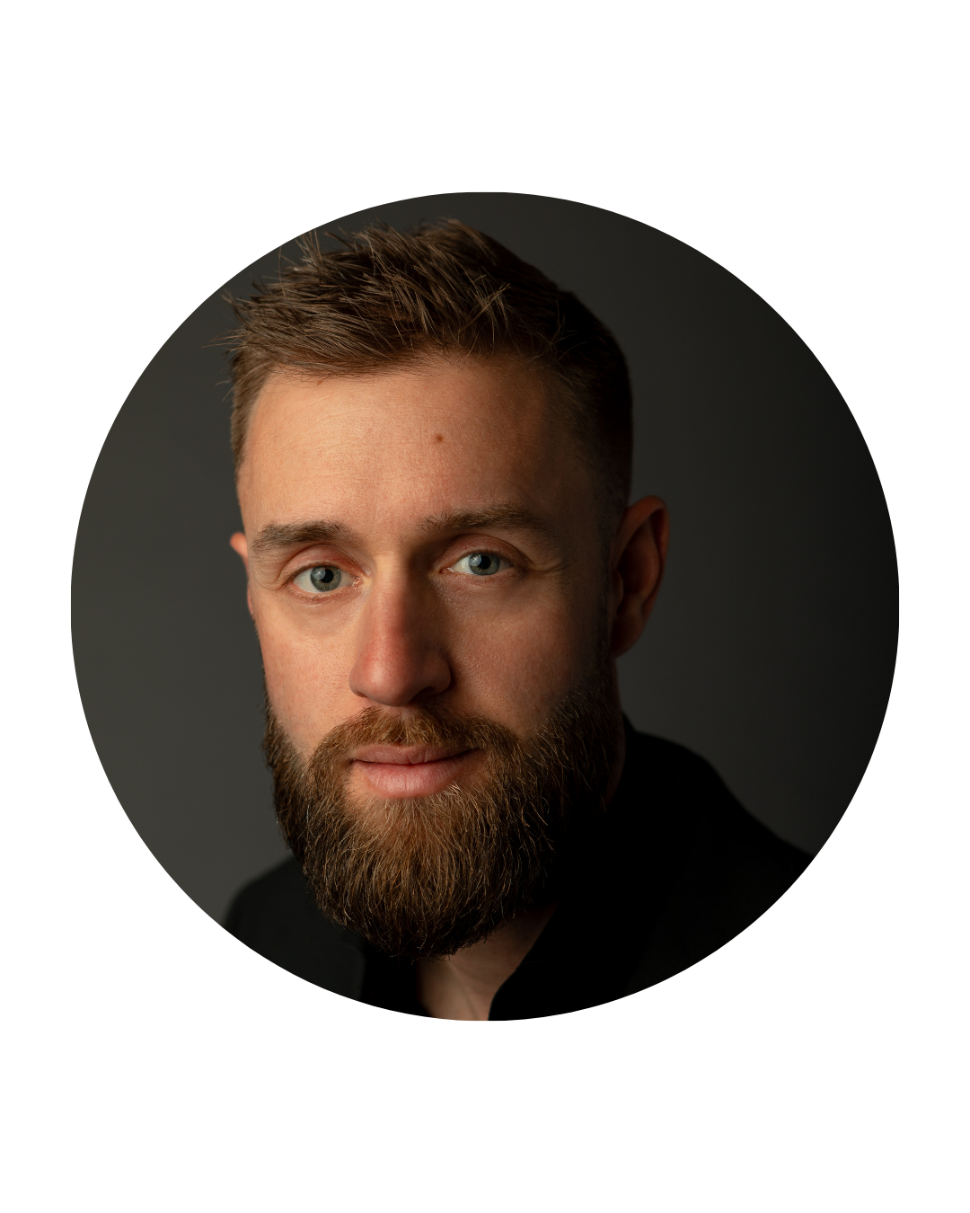 Close-up portrait of a young man with short, styled hair, blue eyes, and a full beard against a dark background.
