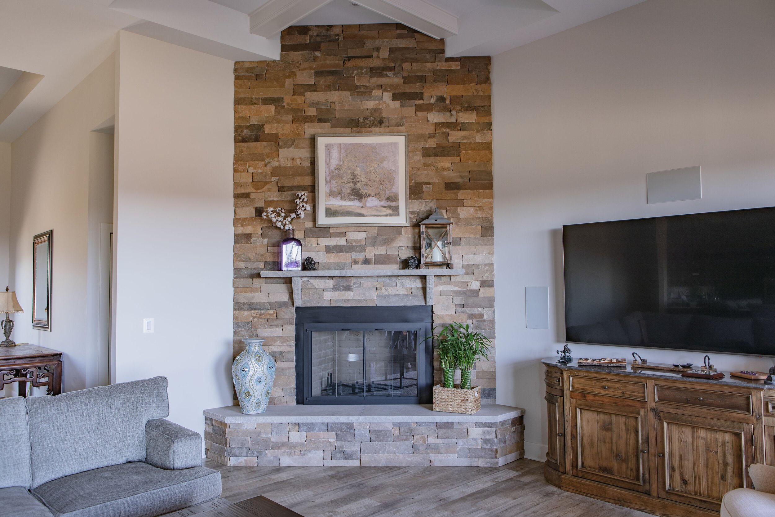 Living room with stone fireplace, framed artwork, decorative vases, houseplants, and furniture including a grey couch and a wooden cabinet.