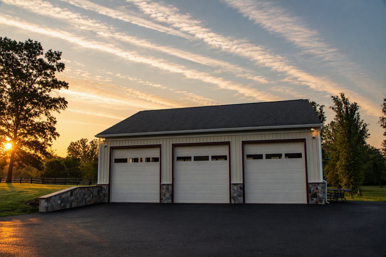 A large garage with three roll-up doors, white with dark trim, situated on a paved driveway at sunset. There is a grassy area with trees and a fence in the background.