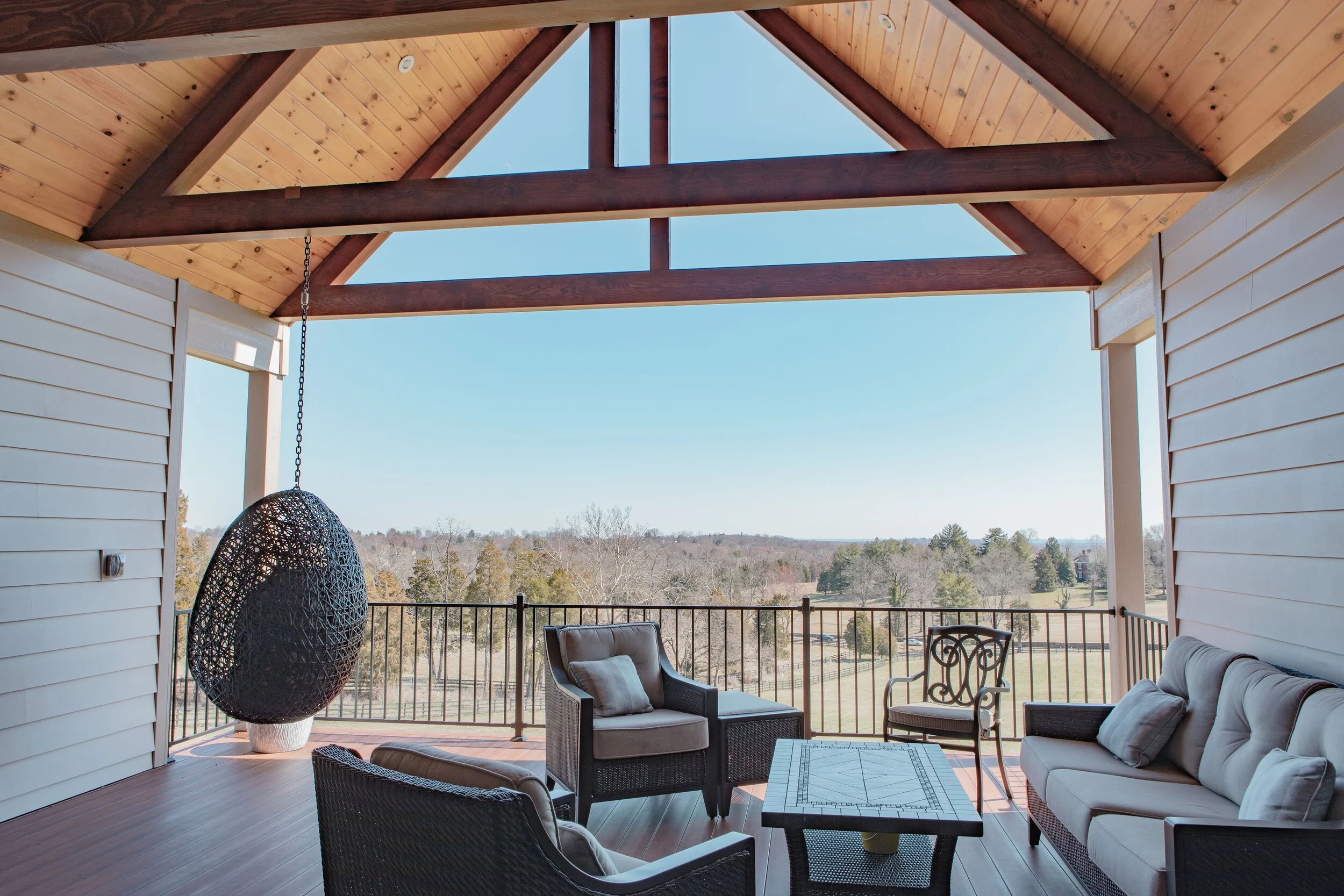 A balcony with outdoor seating, including cushioned chairs and a table, overlooking a scenic landscape with trees and blue sky, and a ceiling with exposed wooden beams.