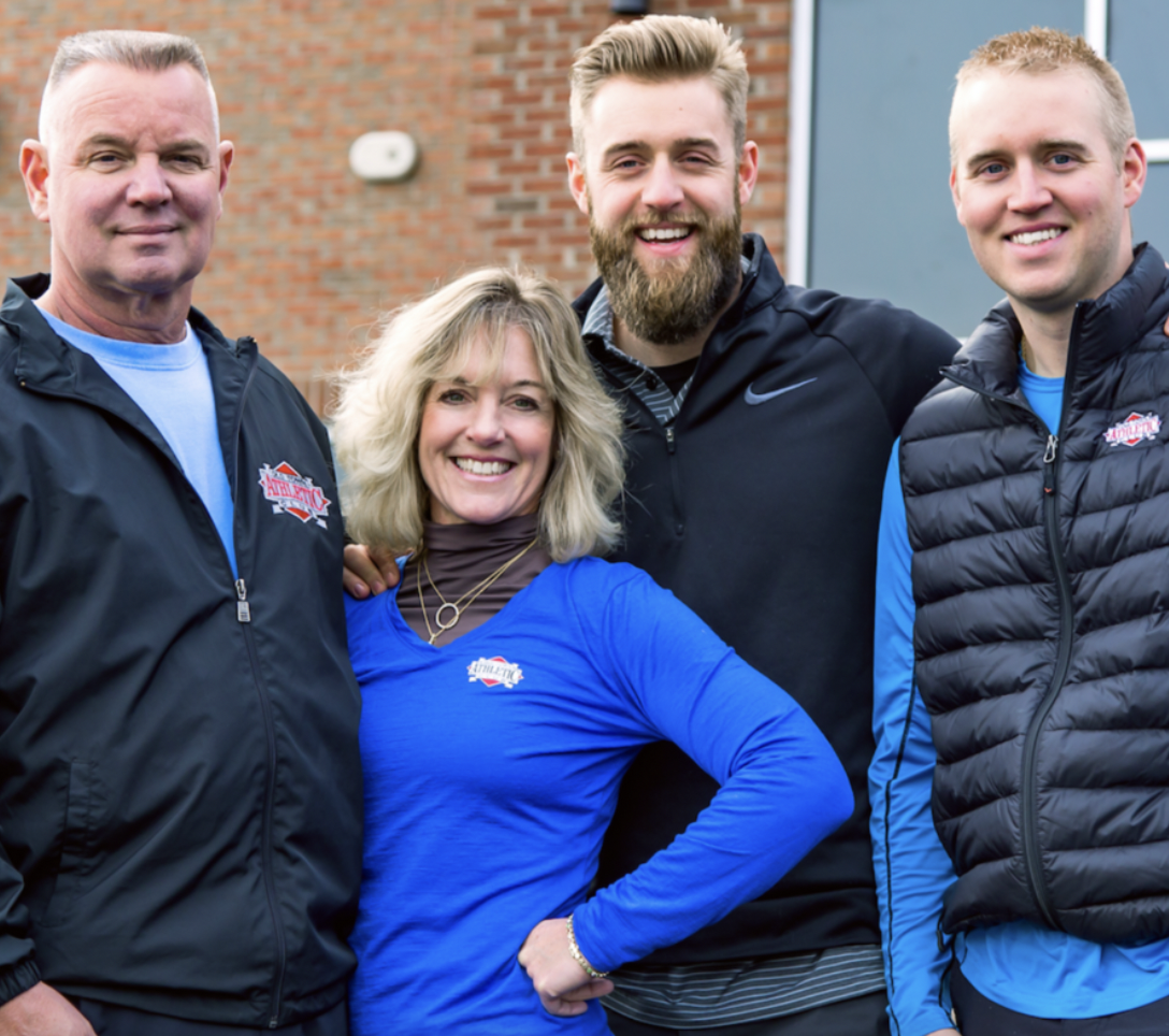Four people standing outdoors, smiling, with a brick building in the background. They are wearing athletic jackets and shirts with athletic team logos.