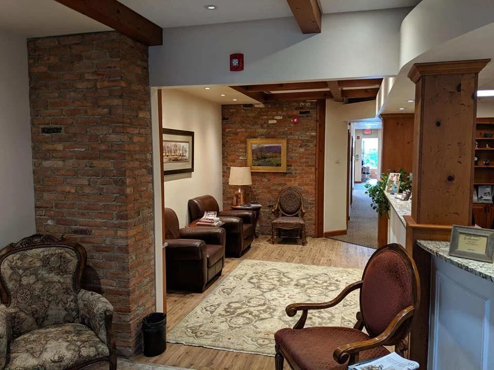 A cozy waiting area with vintage and leather chairs, a brick wall, framed artwork, a floor lamp, and a patterned rug in a warmly lit, rustic interior space.