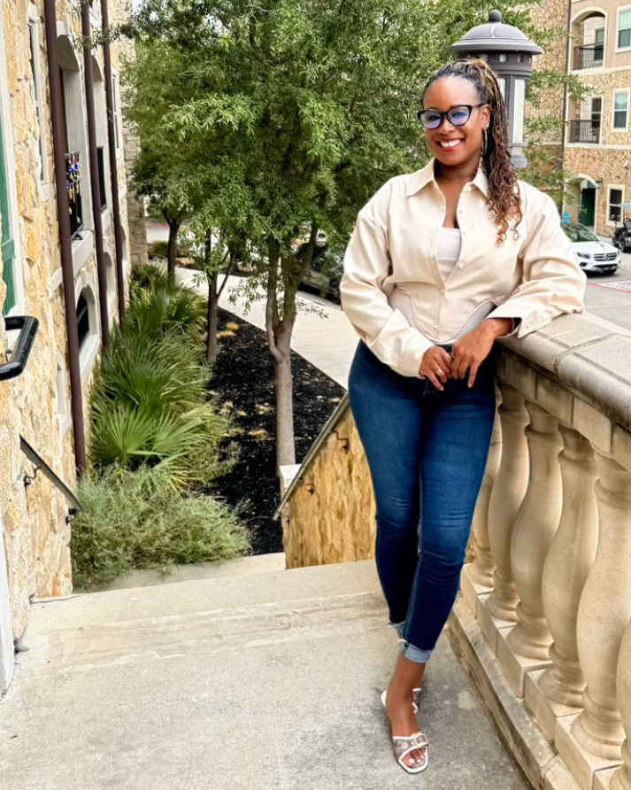 Woman with glasses smiling, standing on an outdoor staircase with a stone railing, greenery and apartment buildings in the background.