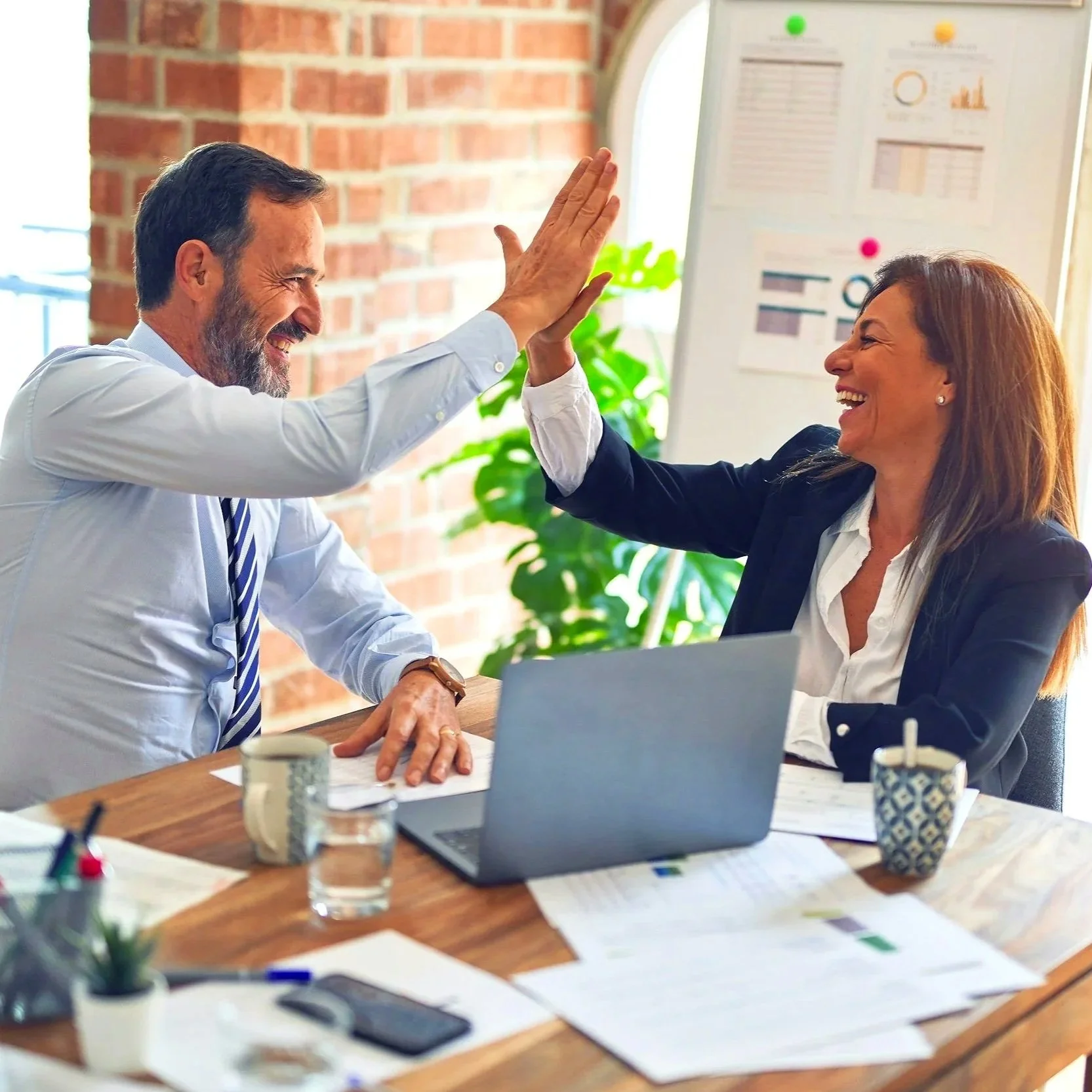 A man and a woman in a business meeting in an office, giving each other a high five and smiling, surrounded by papers, laptops, and coffee cups on a wooden table.