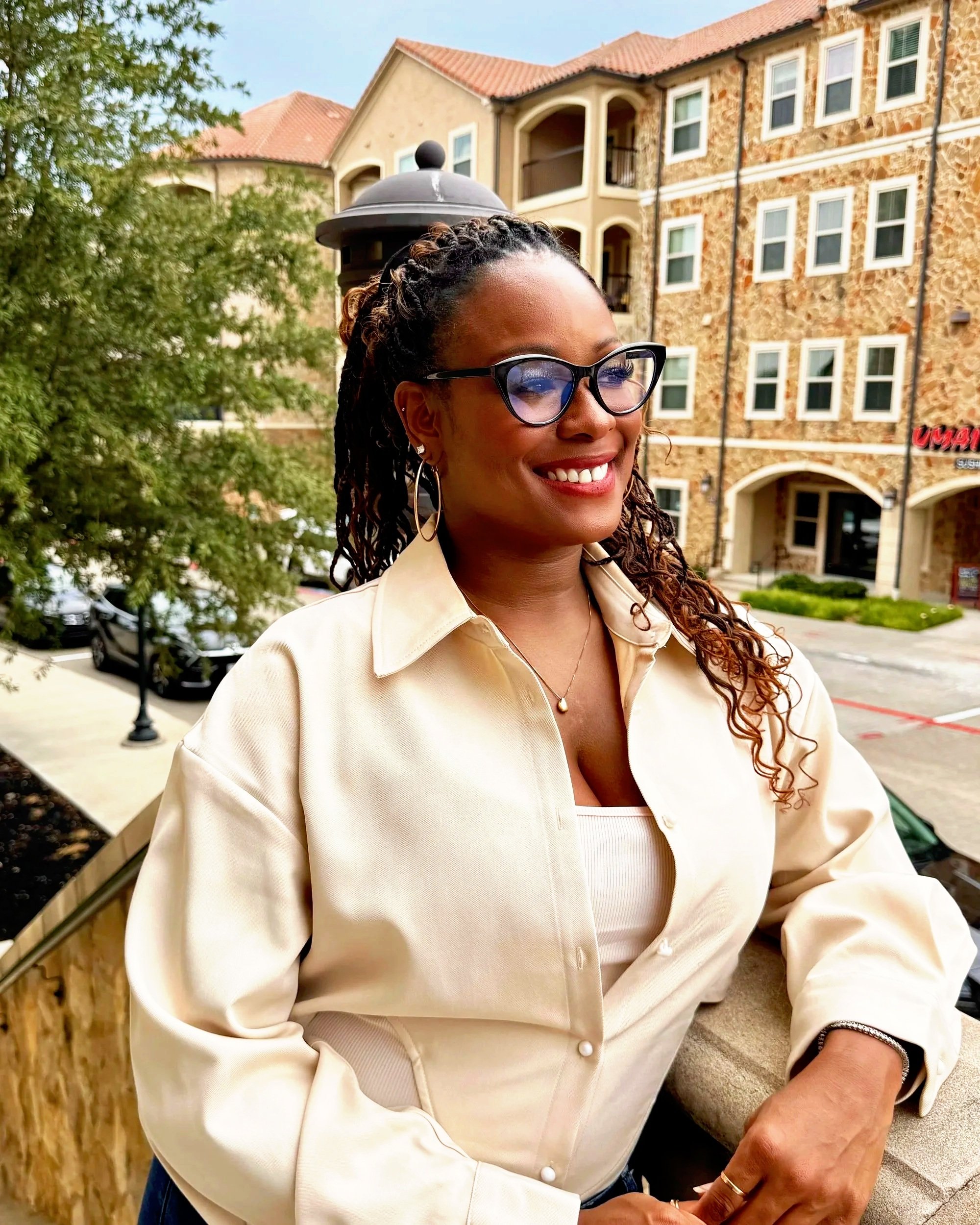 A woman with braids and glasses smiling outdoors with a beige building and trees in the background.