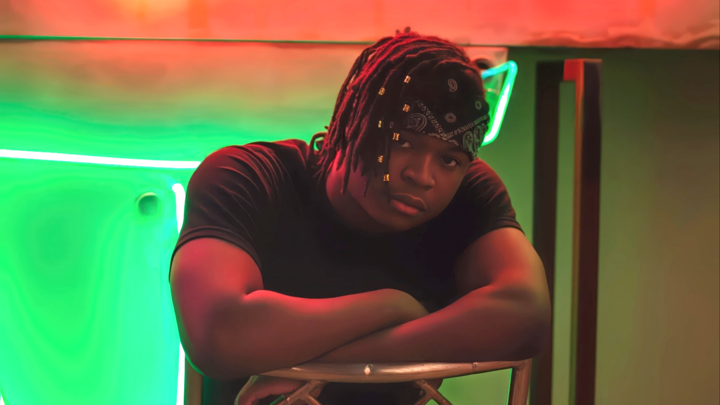 A young man with dreadlocks and a bandana, leaning on a chair with his arms crossed, in front of a neon green and orange background.