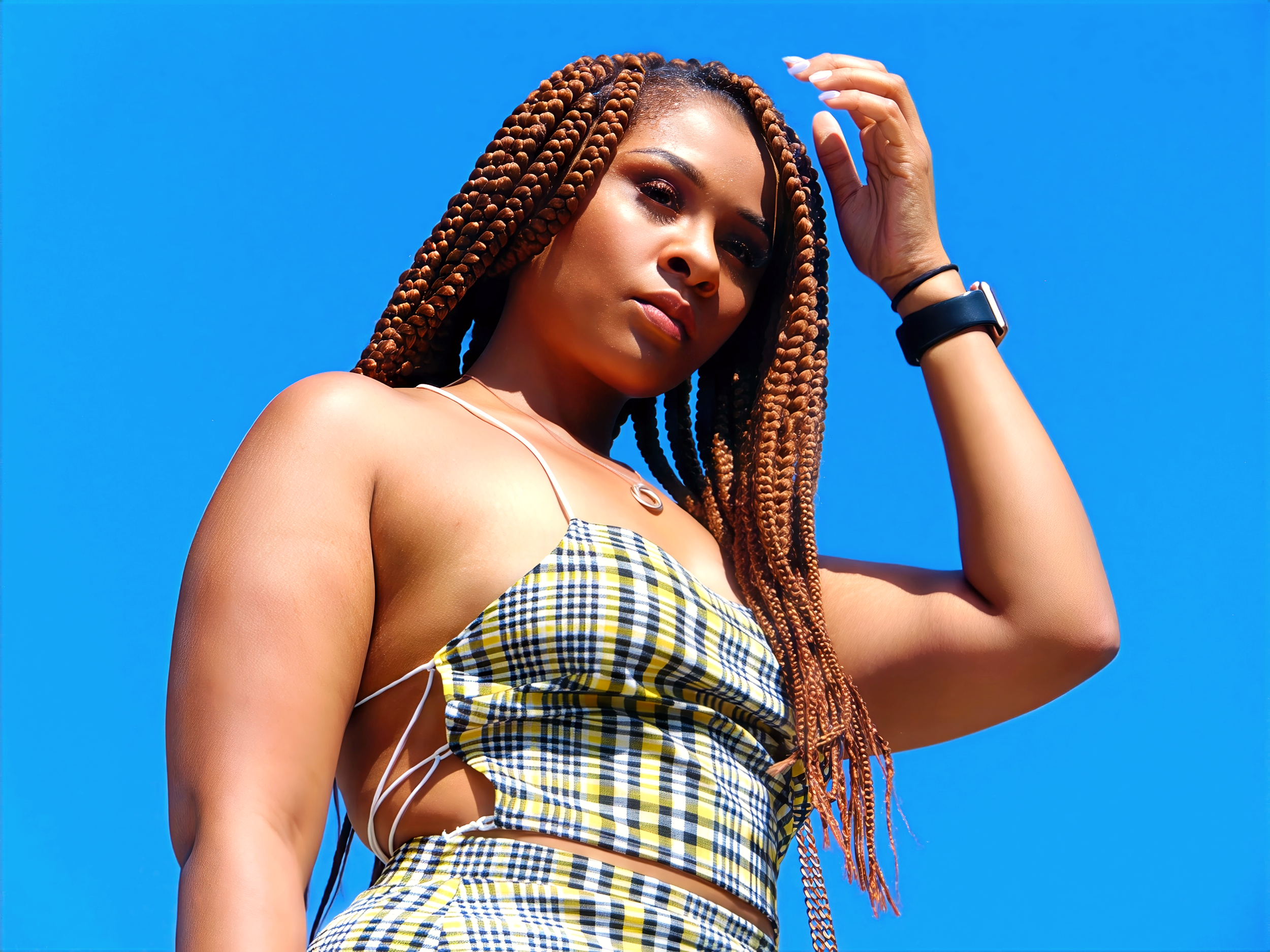 A woman with braided hair and dark skin wearing a yellow and blue checkered top and a black wristband, standing outdoors against a bright blue sky.