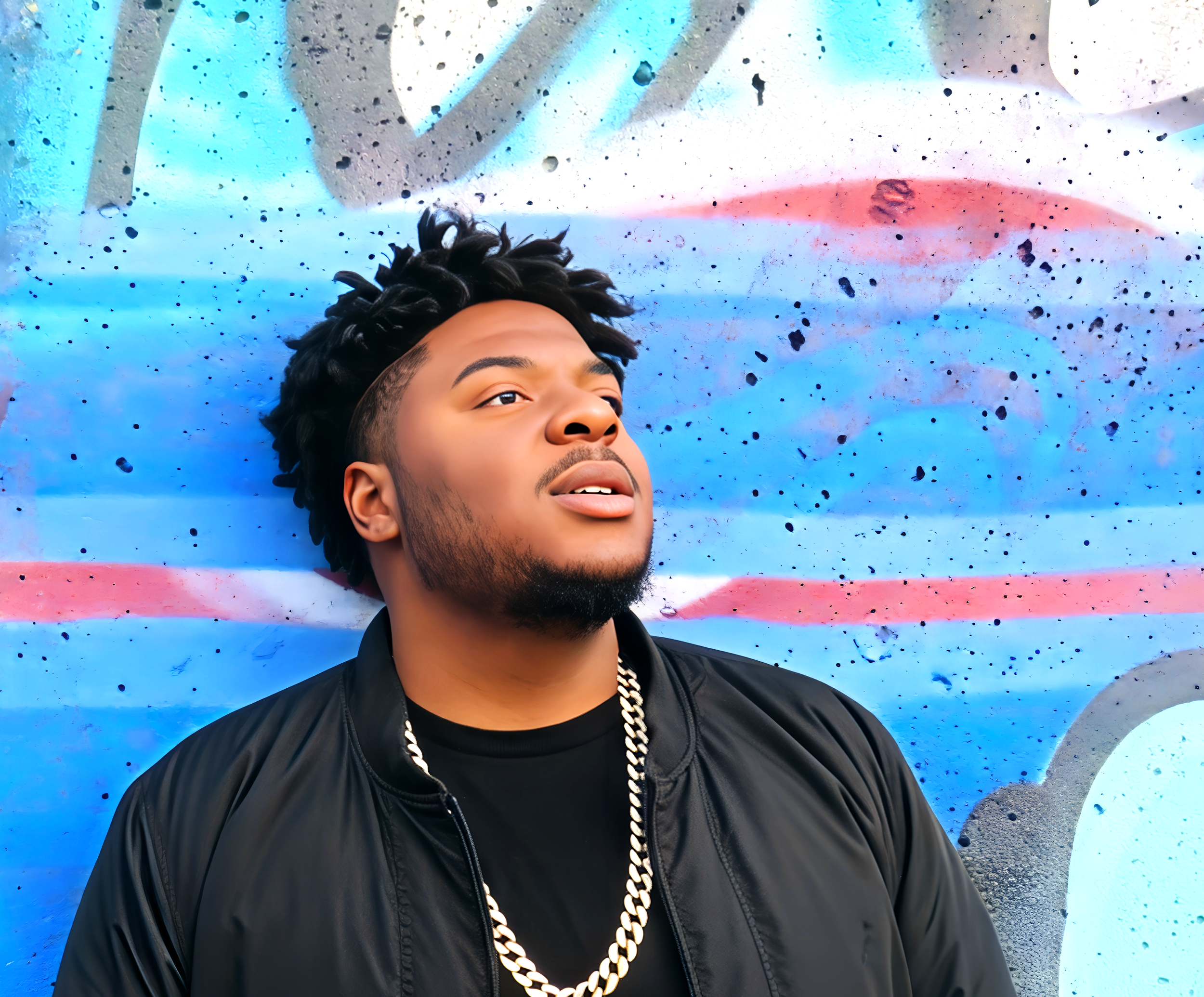 Close-up of a young man with dark, curly hair with highlights, wearing a black leather jacket and a gold chain necklace, lying down in front of a colorful abstract wall with blue, white, and pink paint splatters.