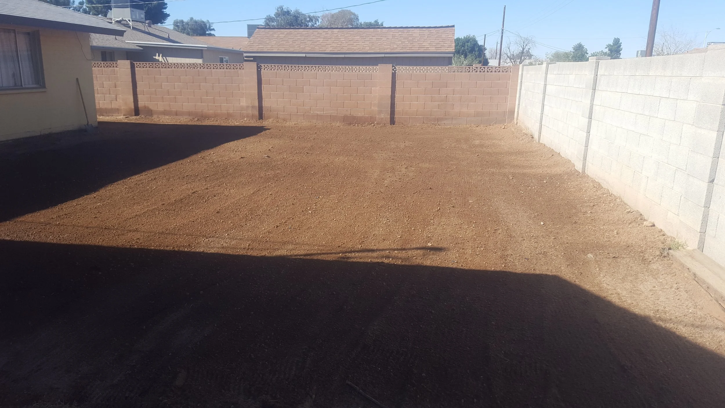 A backyard with a dirt ground, surrounded by a brick wall on the back and a concrete block wall on the right, with part of the house visible on the left.