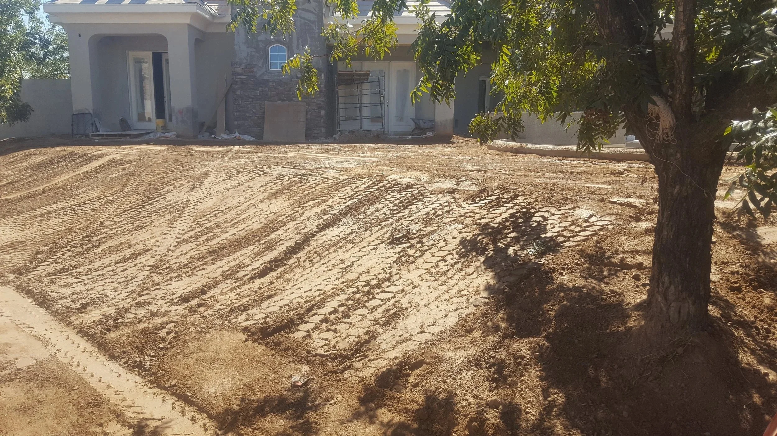 A house under construction with a dirt yard, some construction materials near the walls, and a large tree in the foreground casting shadows.