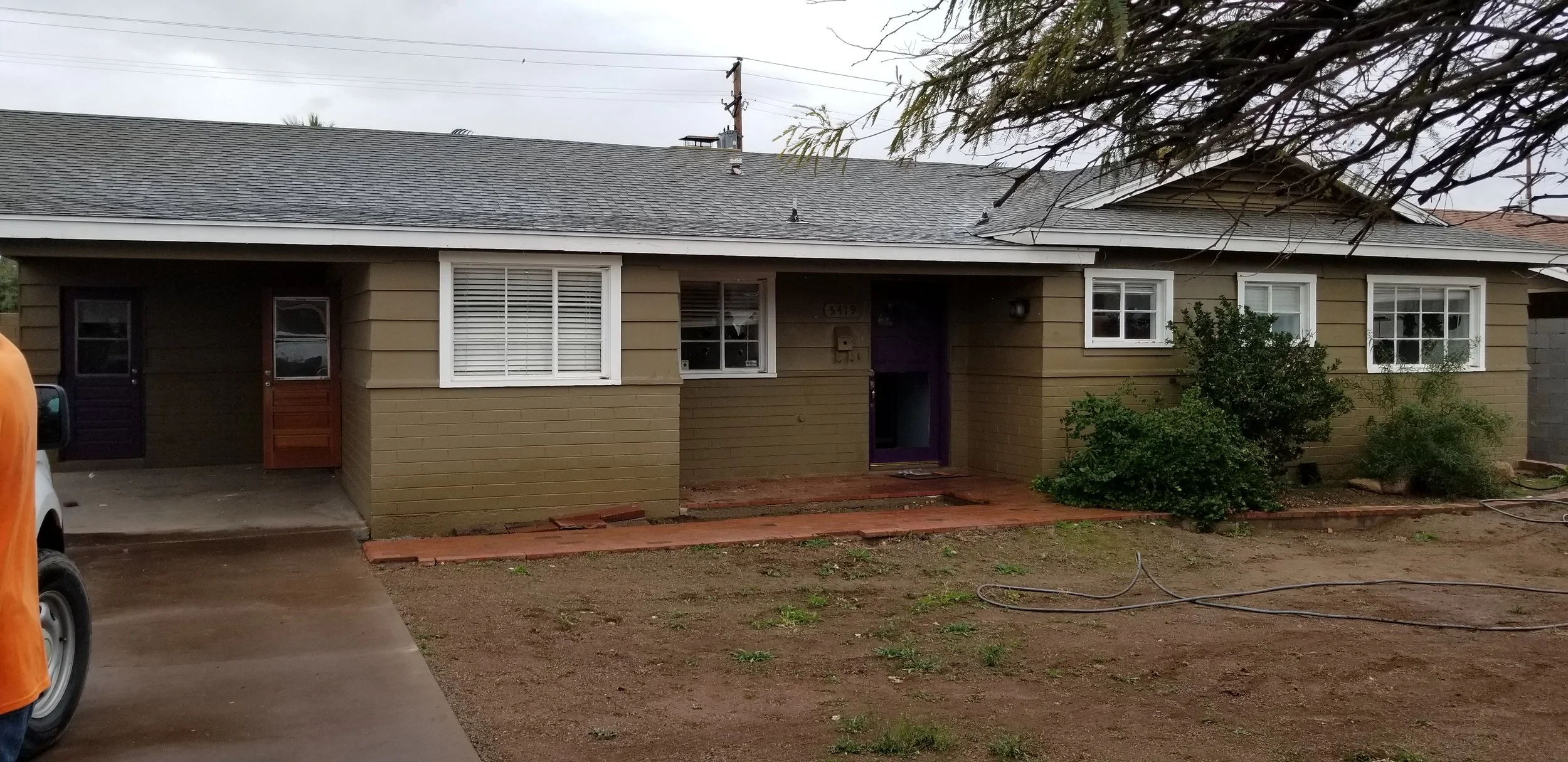 A house with a tan exterior, white window frames, and a purple front door. There are bushes and a tree in front, and the driveway has some construction materials and dirt.