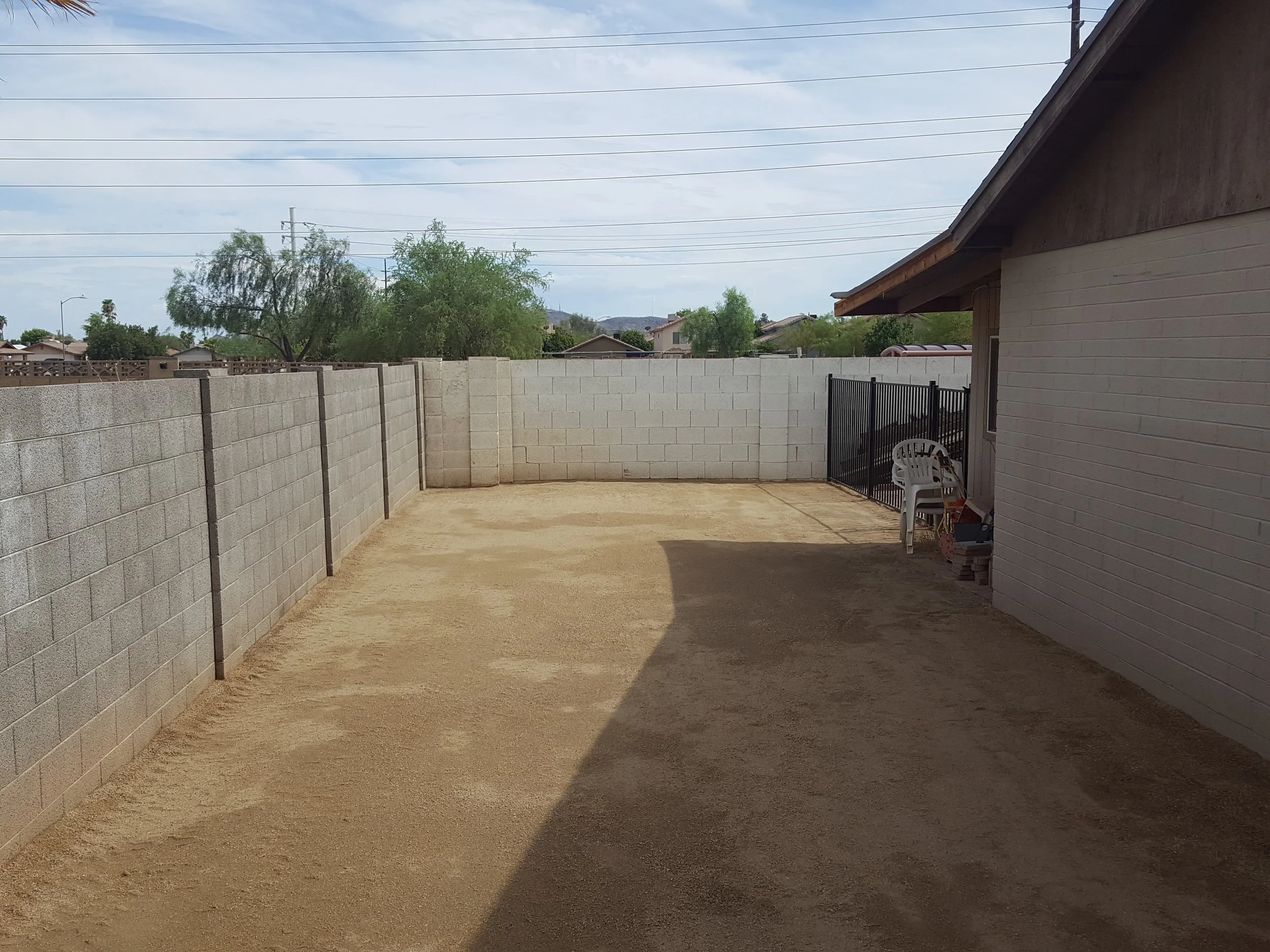 Backyard patio area with concrete block wall, dirt ground, and partial view of house with white brick exterior, black gate, white plastic chairs, and trees in the background under cloudy sky.