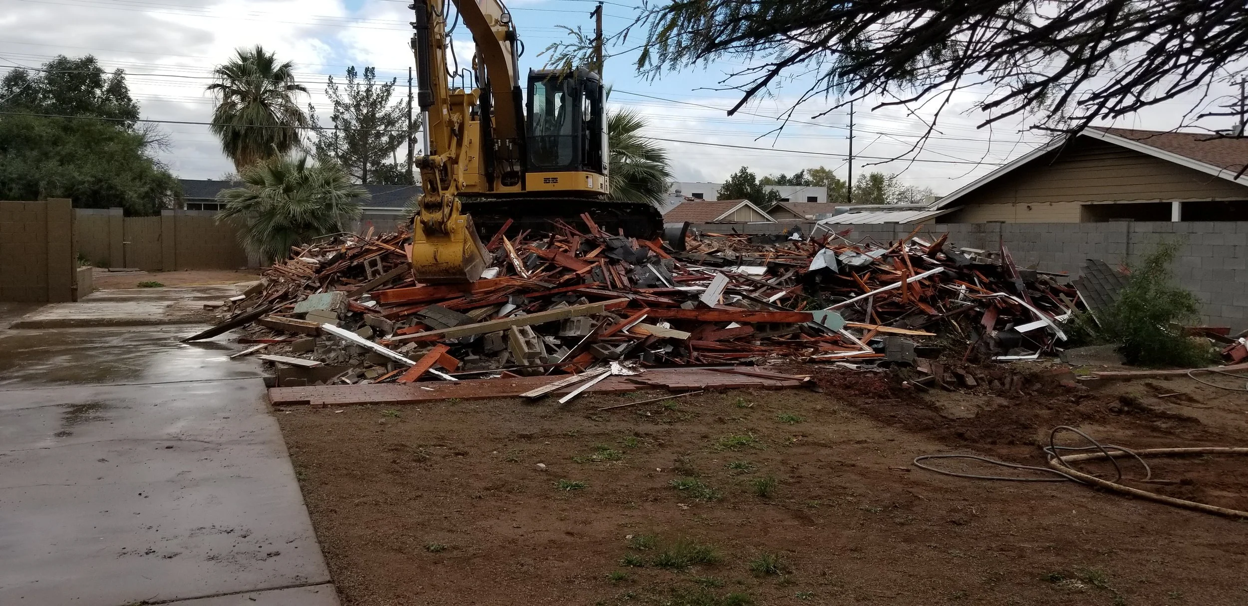 A yellow excavator sitting on a pile of debris in a backyard, with a house, trees, and cloudy sky in the background.