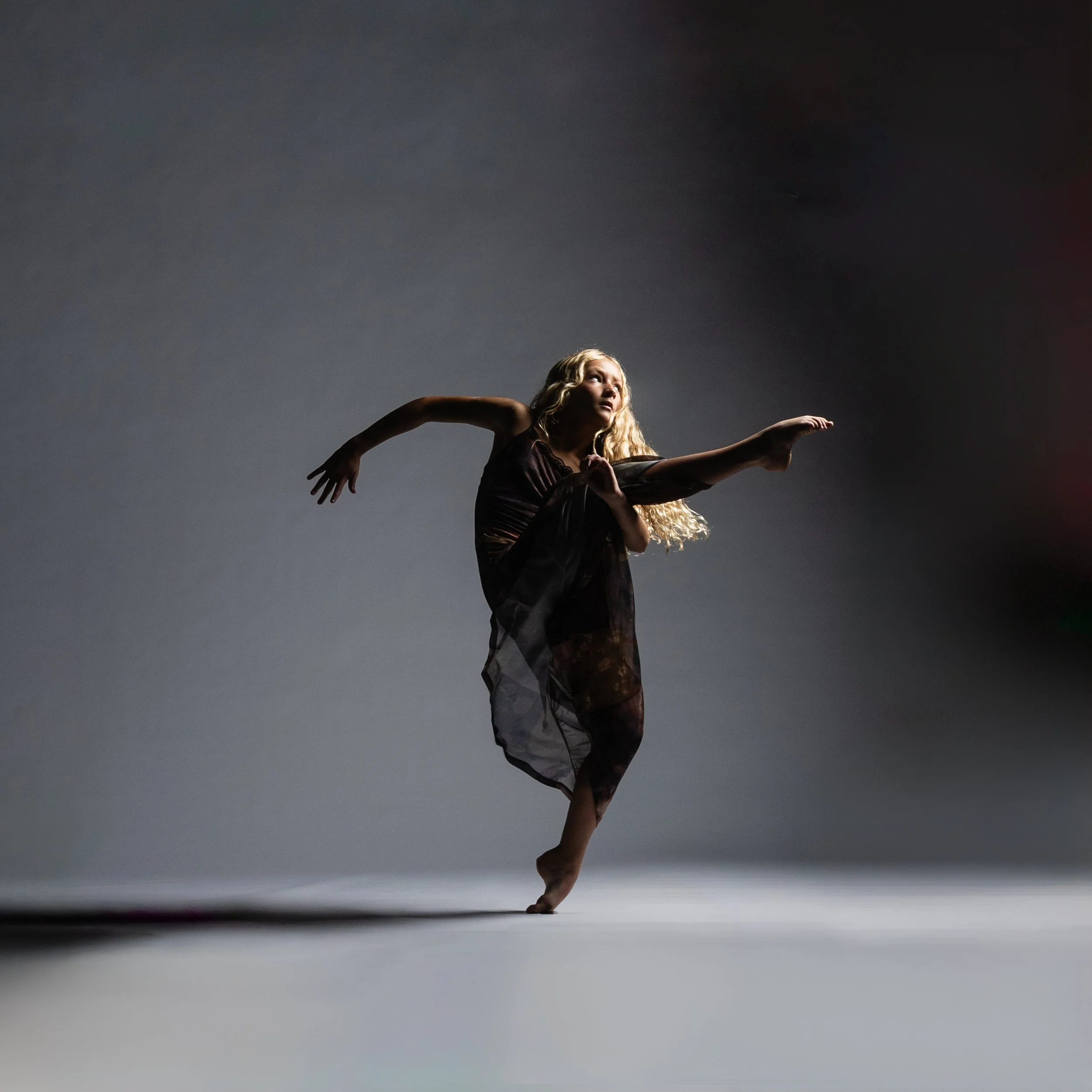 A ballet dancer performing a pose on stage with a dark background, wearing a black costume with a sheer skirt.