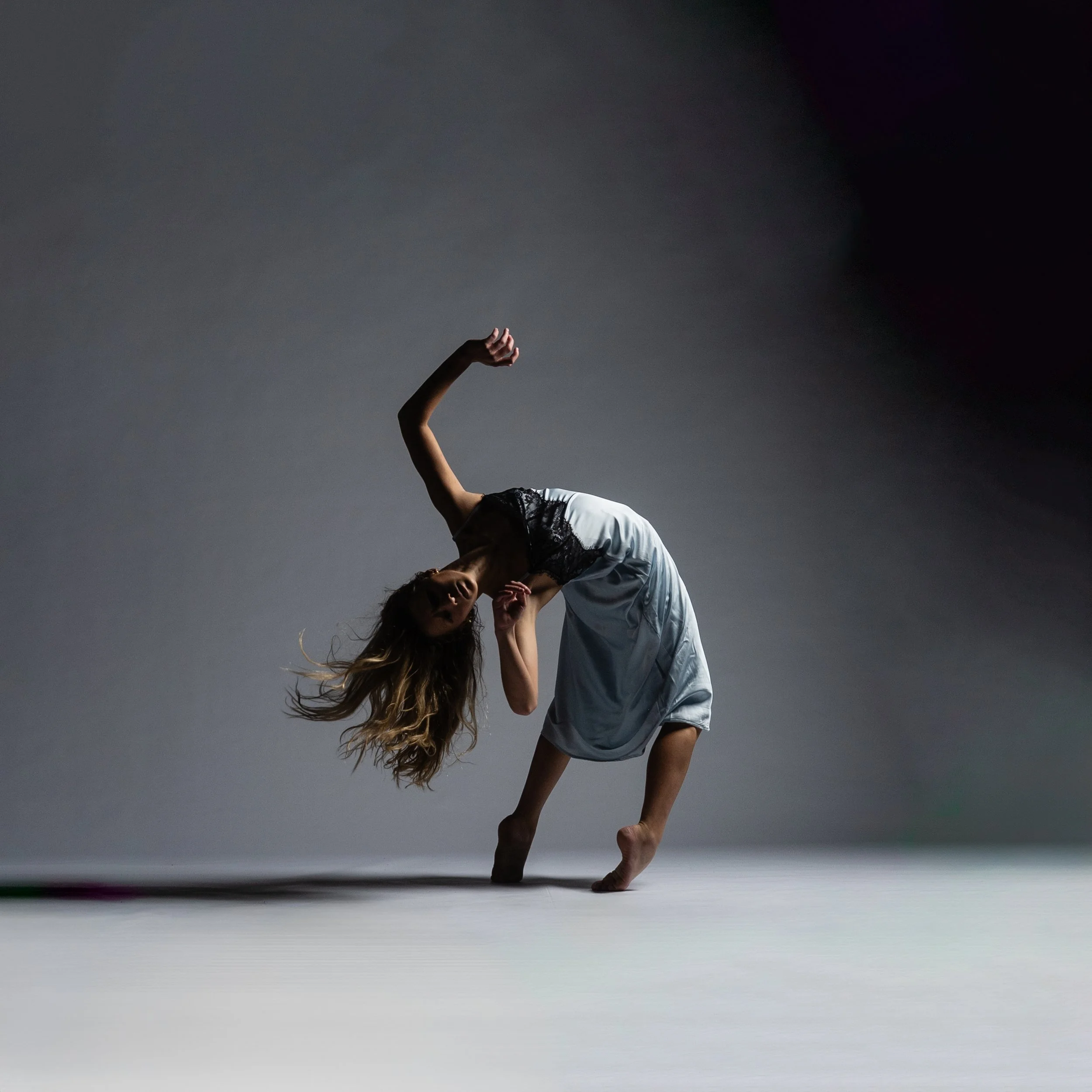 A person with long hair performing a handstand with one arm on the ground in a studio with a dark background.