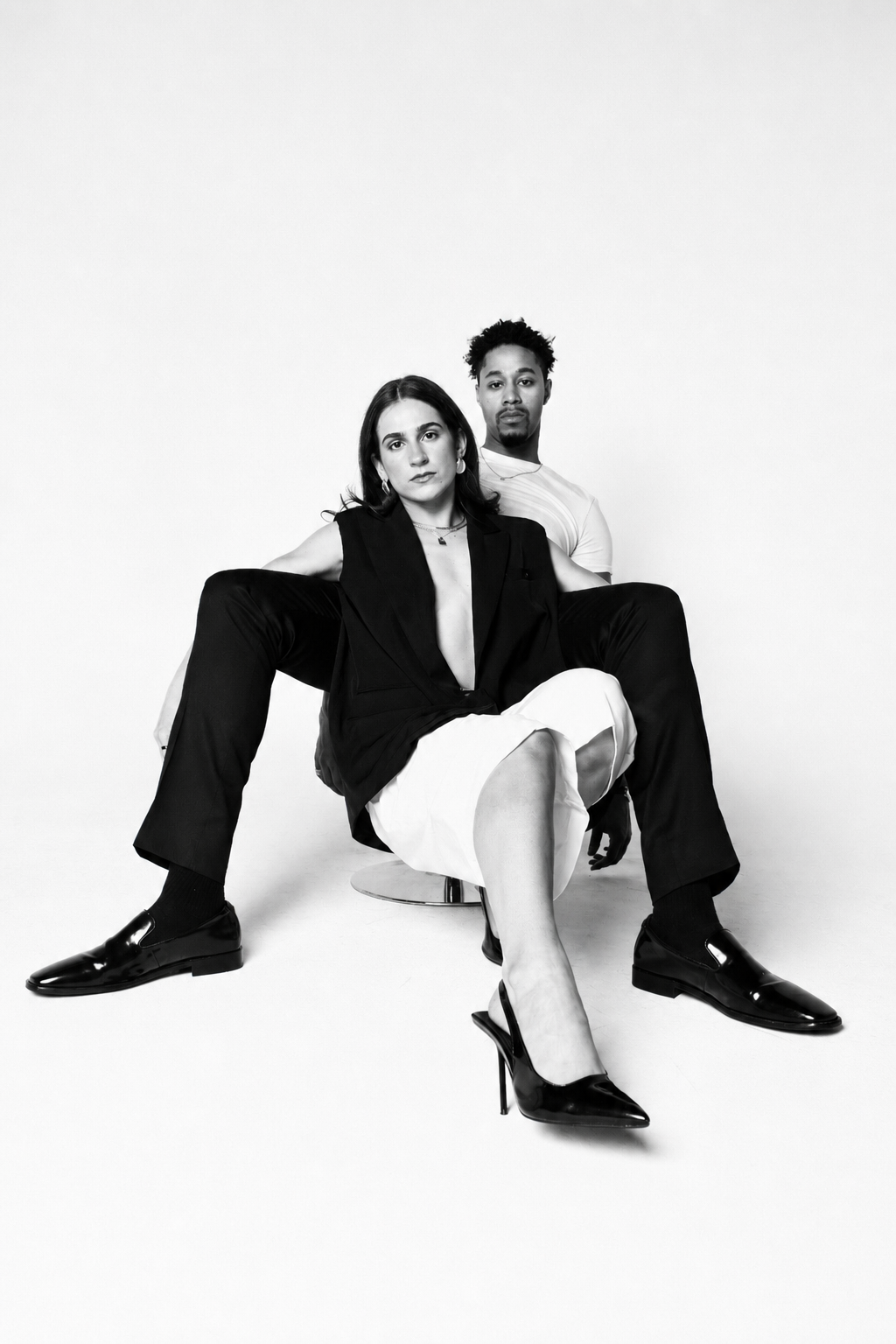 Black and white photo of a woman and man posing together, with the woman seated on a stool and the man sitting behind her on the floor, against a plain white background.
