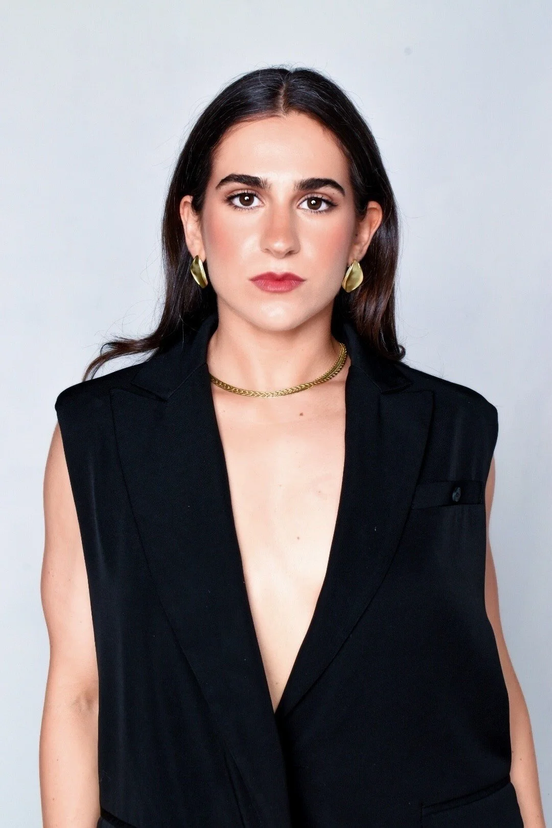A woman with dark hair and brown eyes wearing a sleeveless black blazer with gold jewelry against a plain light background.