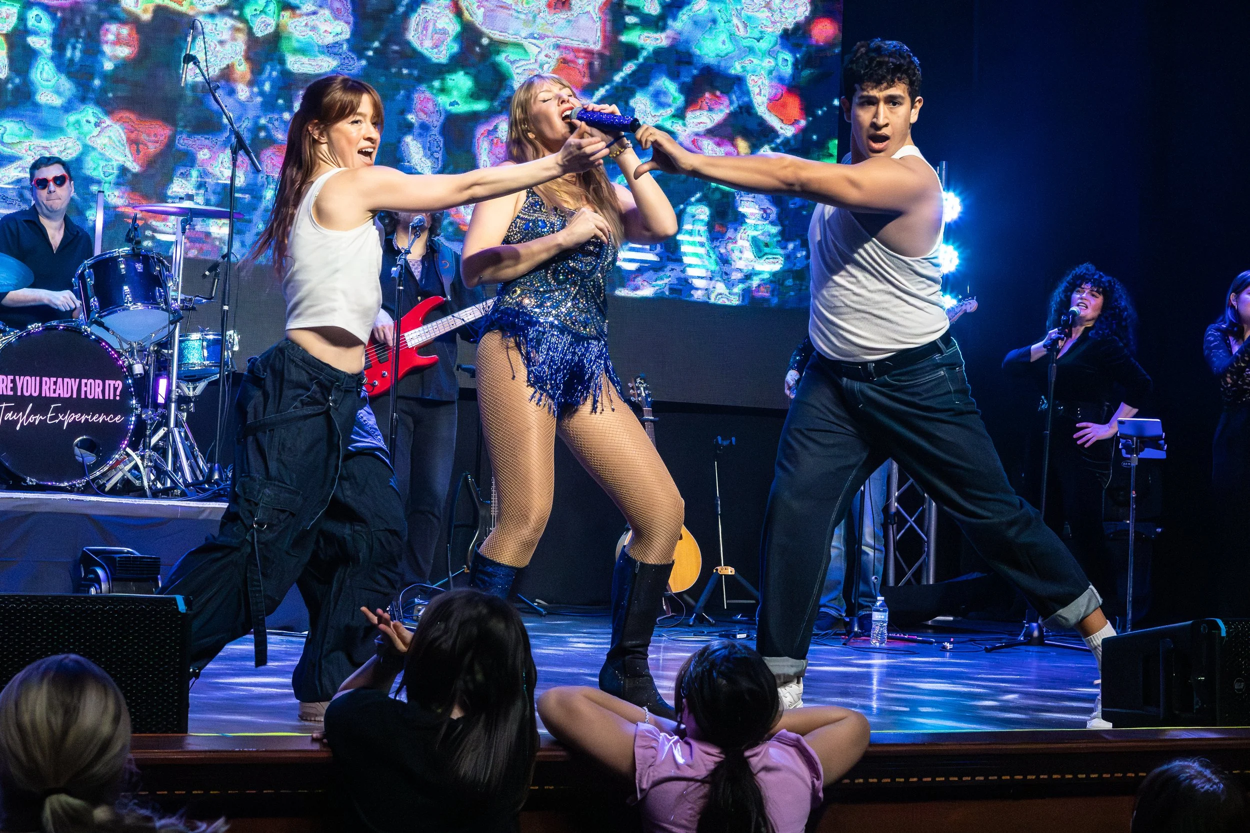 A live musical performance featuring a group of young people on stage. One woman in fishnet stockings and a sparkly top is singing into a microphone held by a man. Two women are dancing, and a drummer and guitarist are visible in the background. Audience members are seen in the foreground, watching the performance.