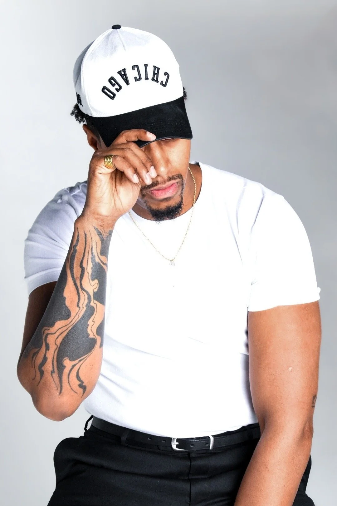 A man wearing a black and white cap with the word 'CELEBRIDO', a white t-shirt, and black pants, touching his hat while looking down, with tattoos on his right arm, a gold ring on his finger, and a delicate chain necklace.