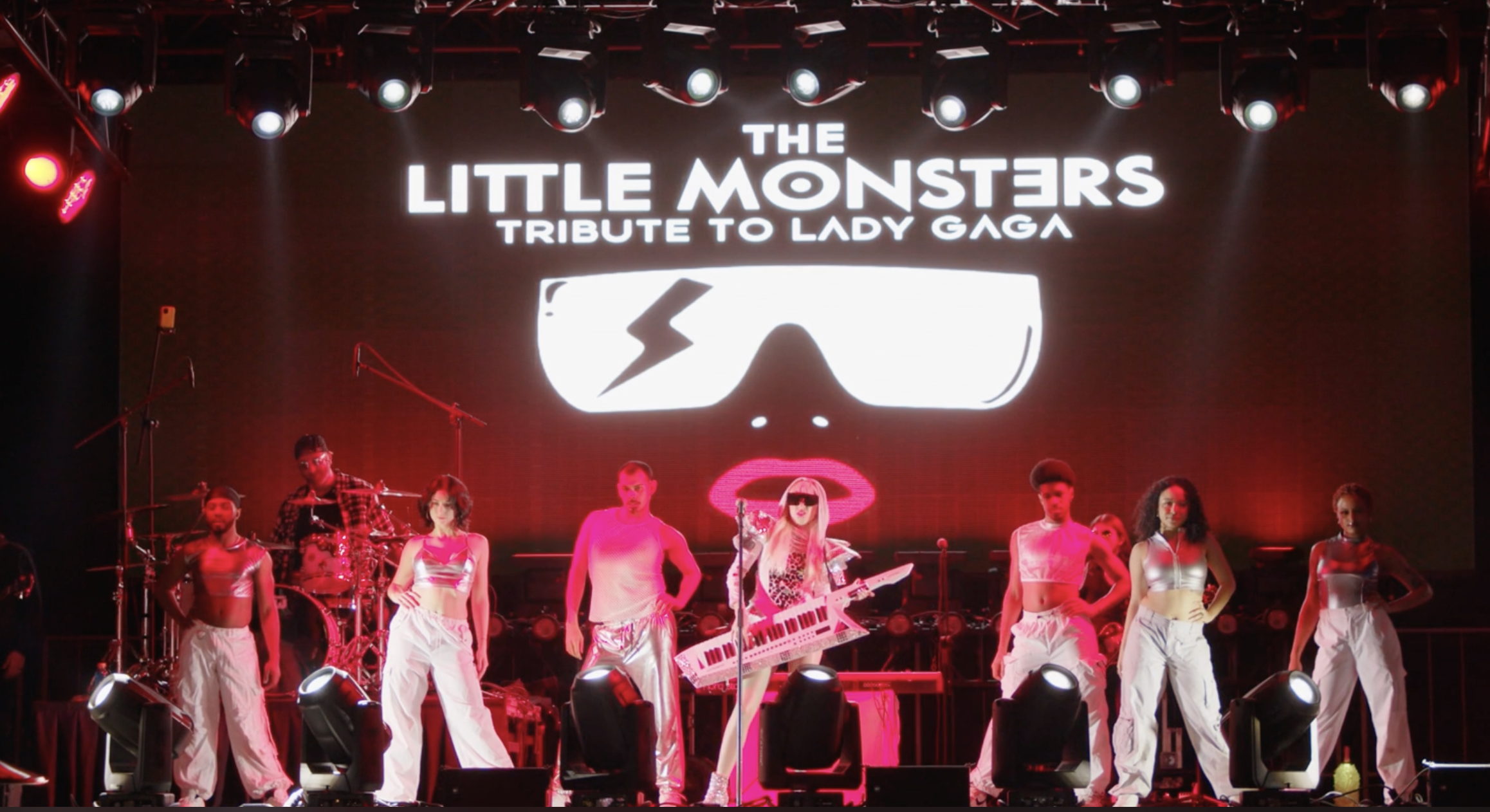 Stage performance featuring Lady Gaga tribute artist with a keyboard, accompanied by dancers in white outfits, with a large screen above displaying text about Little Monsters tribute to Lady Gaga in bright neon lights.