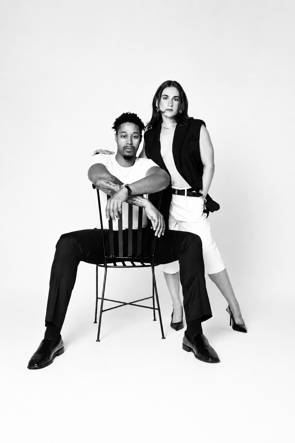 Black and white photo of a man sitting on a chair and a woman standing next to him against a plain background. The man has tattoos on his arms and is wearing a white t-shirt, black pants, and black shoes. The woman has long dark hair, is wearing a sleeveless blazer, white knee-length pants, and high heels. She has her arm on the man's shoulder and looks directly at the camera.