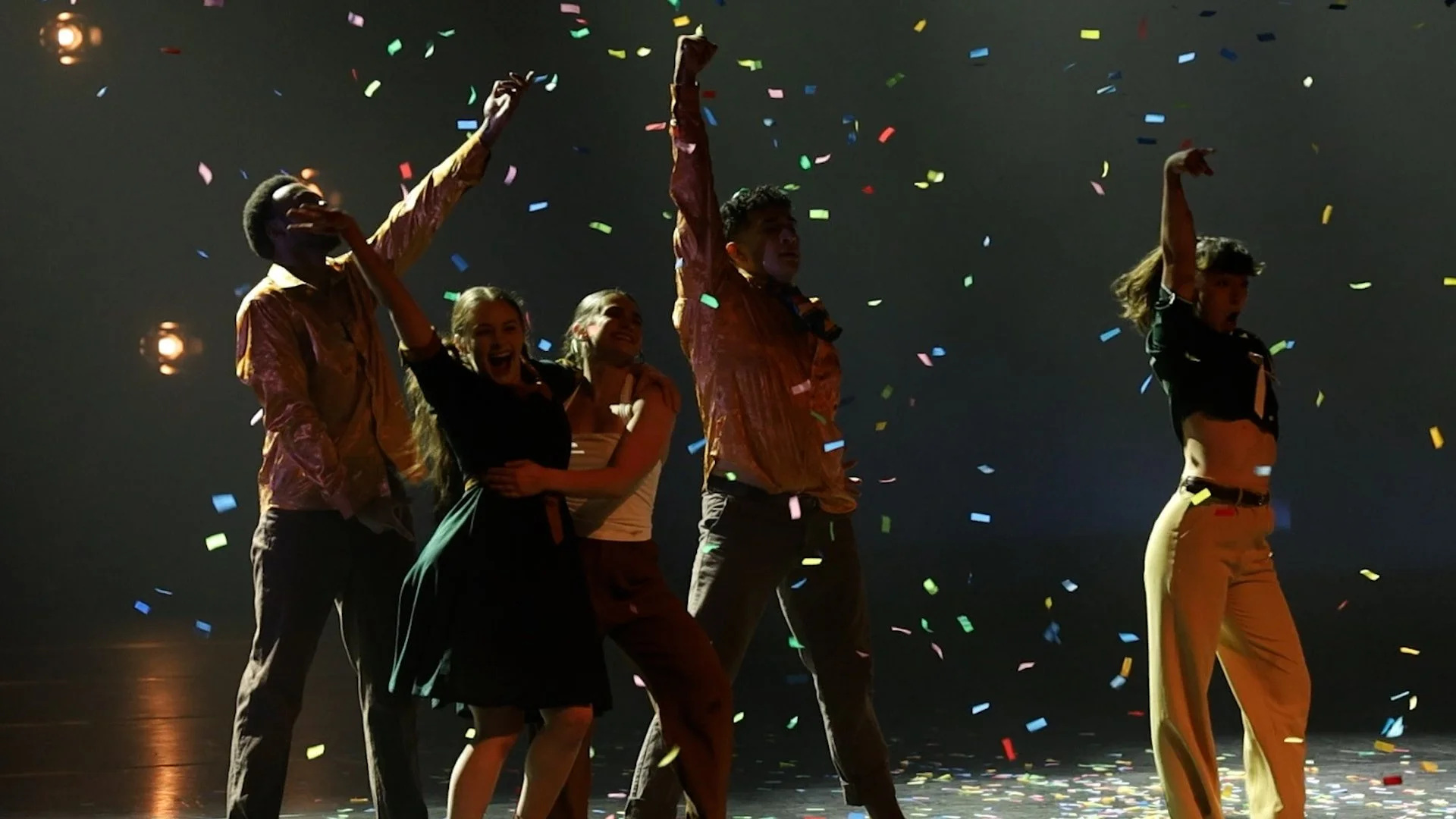 A group of five young people celebrating on stage with colorful confetti falling around them, some raising their arms in victory or joy.