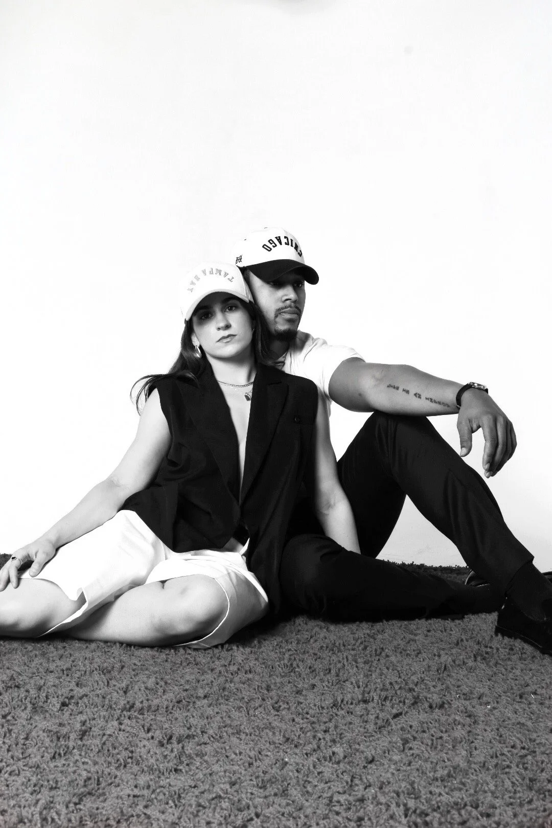 Black and white photo of a woman and a man sitting on a carpeted floor against a plain background. The woman is sitting cross-legged, wearing a sleeveless vest, white shorts, and a cap. The man is sitting with his legs bent, wearing a T-shirt, dark pants, and a cap. They both have serious expressions.
