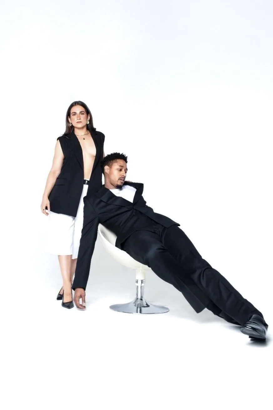 A woman and a man posing in studio with a white background. The woman stands while the man leans against a chair, dressed in fashionable black and white clothing.