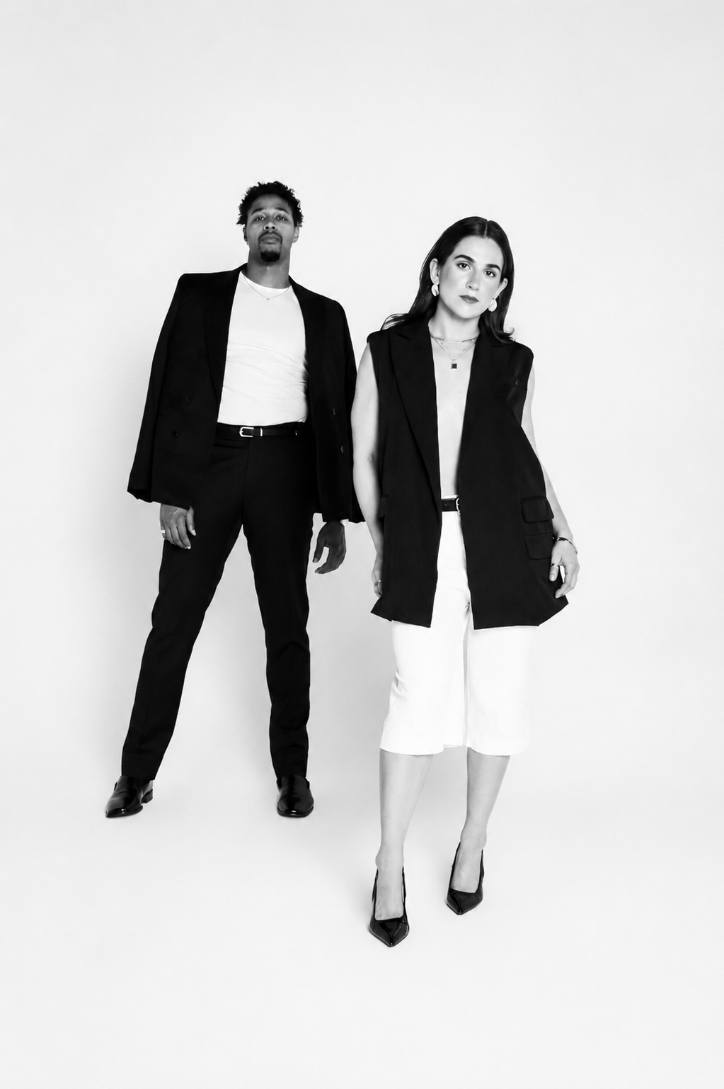 Two stylish people, a man and a woman, standing against a plain white background. The man is wearing a black blazer, white shirt, and black pants. The woman is dressed in a sleeveless black vest, white pants, and high heels.