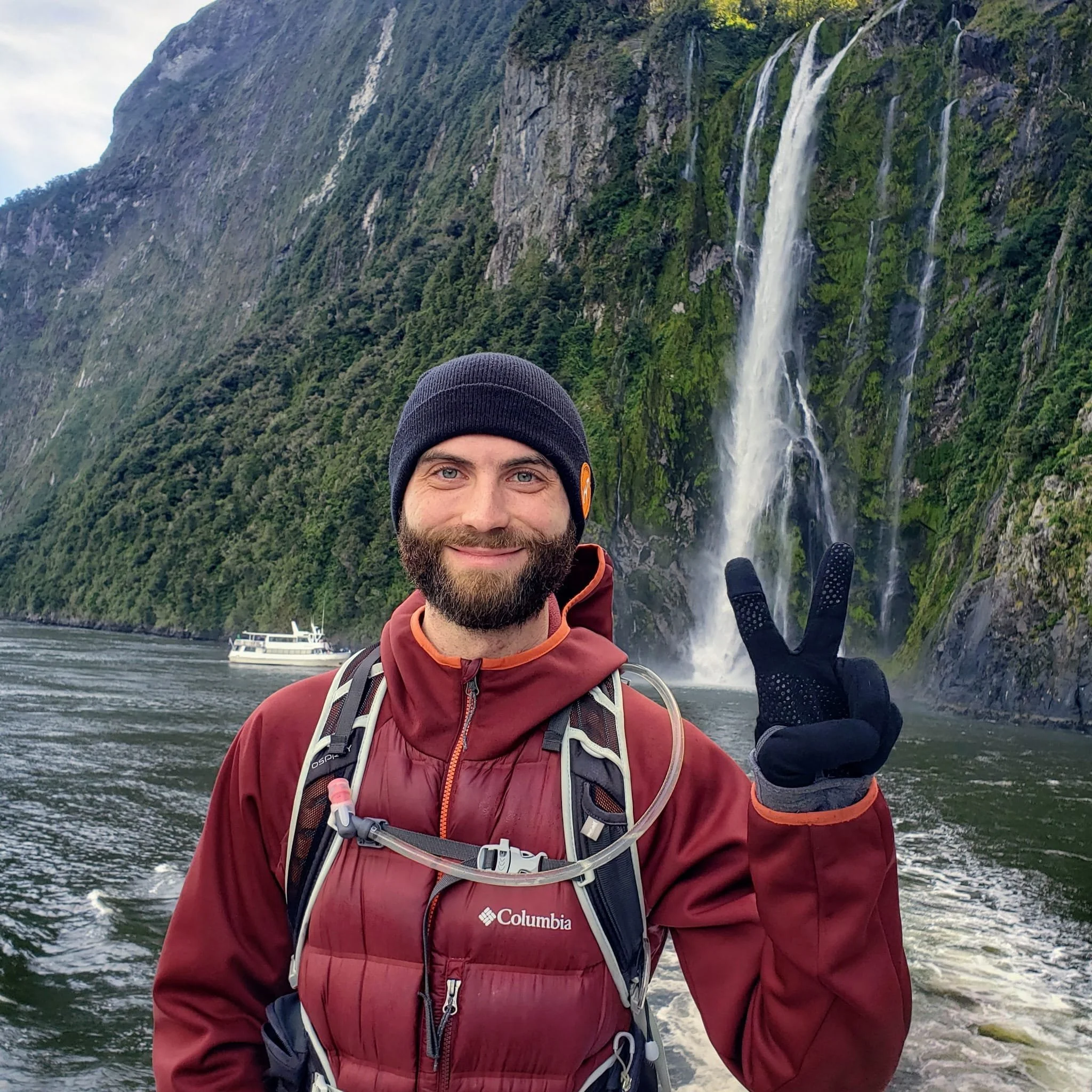 A man with a beard and blue eyes wearing a black beanie, red Columbia jacket, and a backpack, smiling at the camera, standing in front of a waterfall with lush green forest on a hillside and a boat in the water nearby.