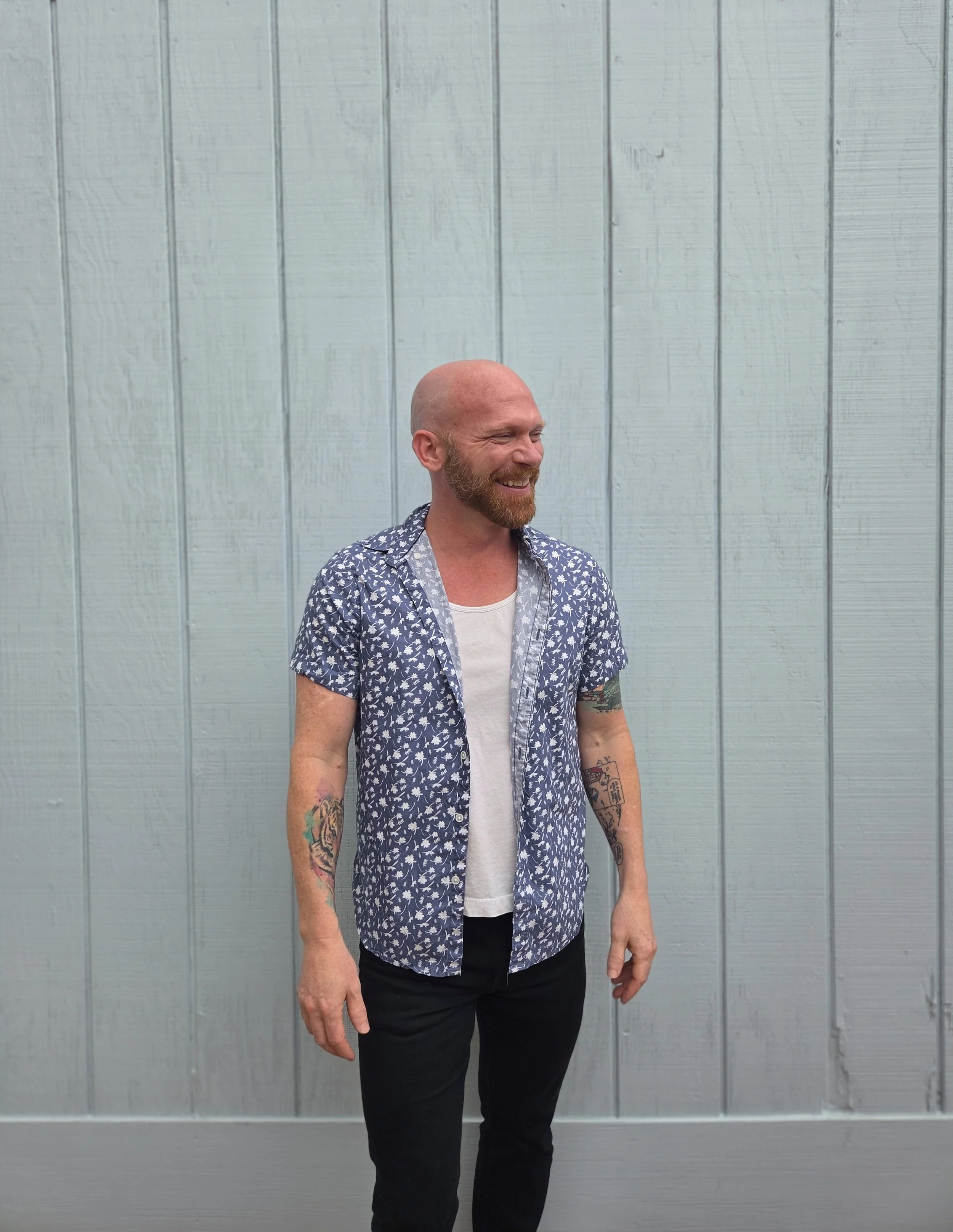 A smiling, bald man with a beard wearing a short-sleeved, button-up shirt with a floral pattern, a white T-shirt underneath, and black pants standing in front of a light blue wooden wall.