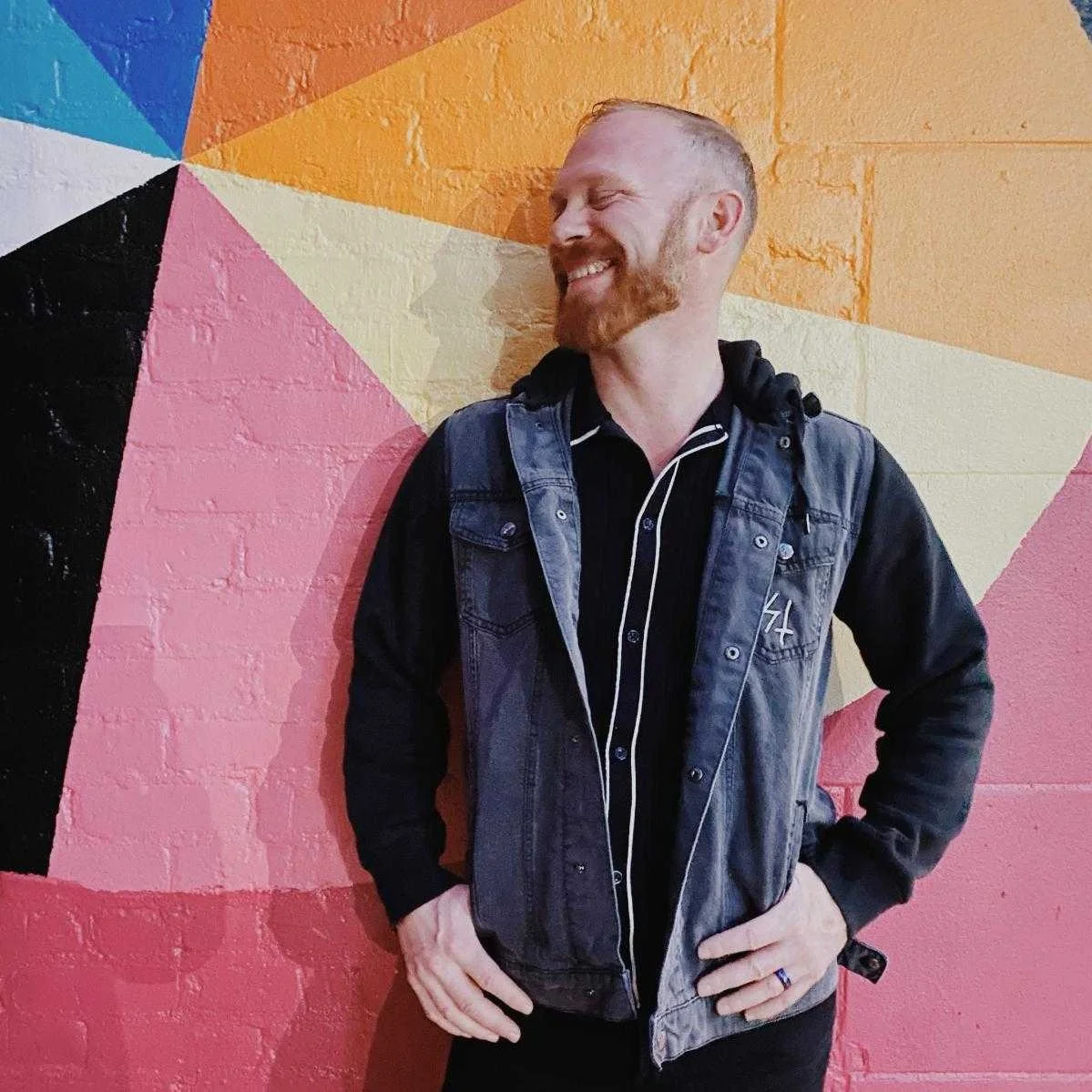 A smiling man with a beard and short hair standing against a colorful geometric mural wall with pink, black, yellow, orange, blue, and white shapes. He is wearing a black hoodie under a denim vest.