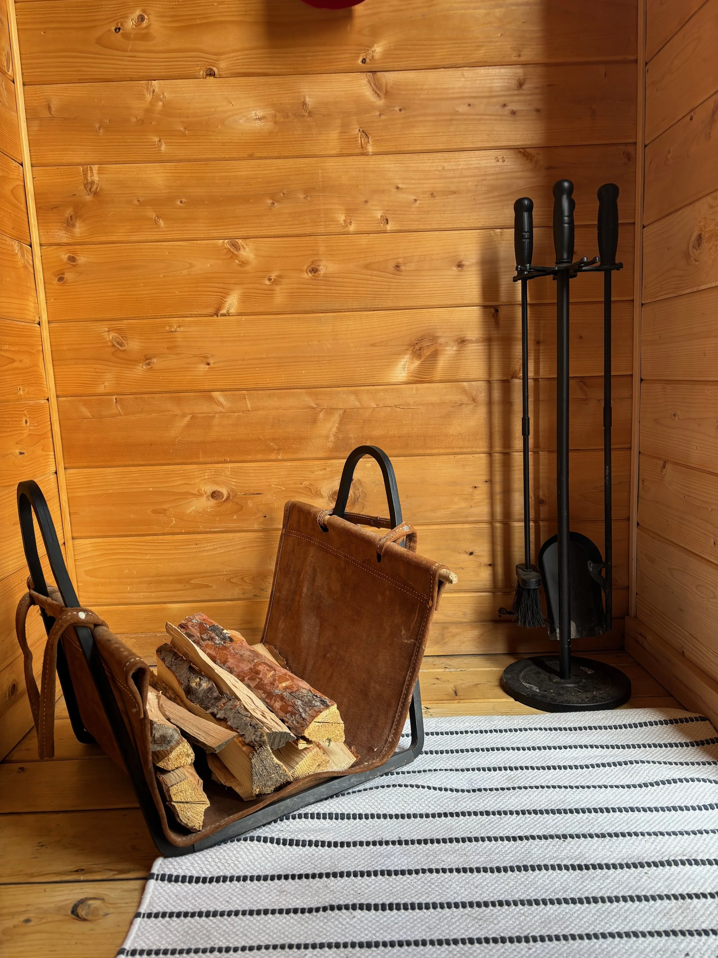 Wood-paneled room with a metal log holder filled with chopped firewood, a striped rug on a wooden floor, and a black fireplace tool set on a stand.