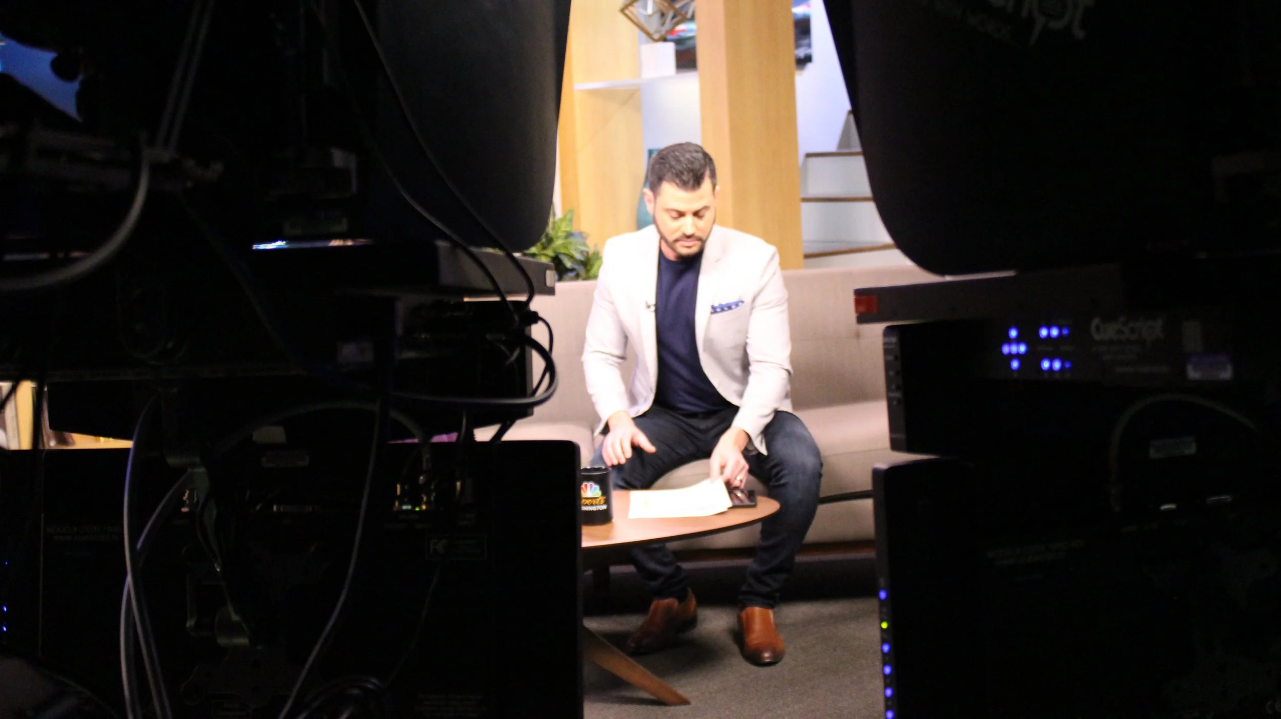 A man in a white blazer and dark pants sitting on a beige couch, looking down at a notebook or document on his lap, with a coffee mug on a small table in front of him, surrounded by studio equipment and cameras in a television or video recording studio.