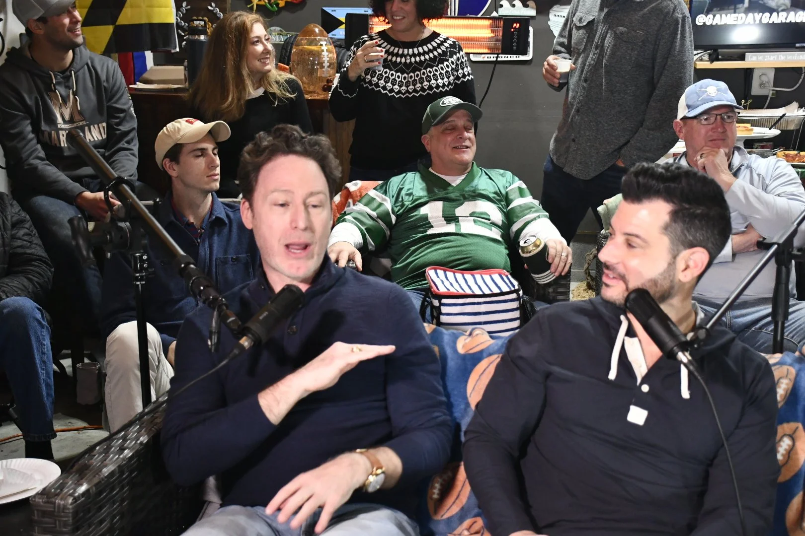 A group of people gathered indoors, some sitting and some standing, engaging in conversation and enjoying drinks, with microphones and recording equipment present.