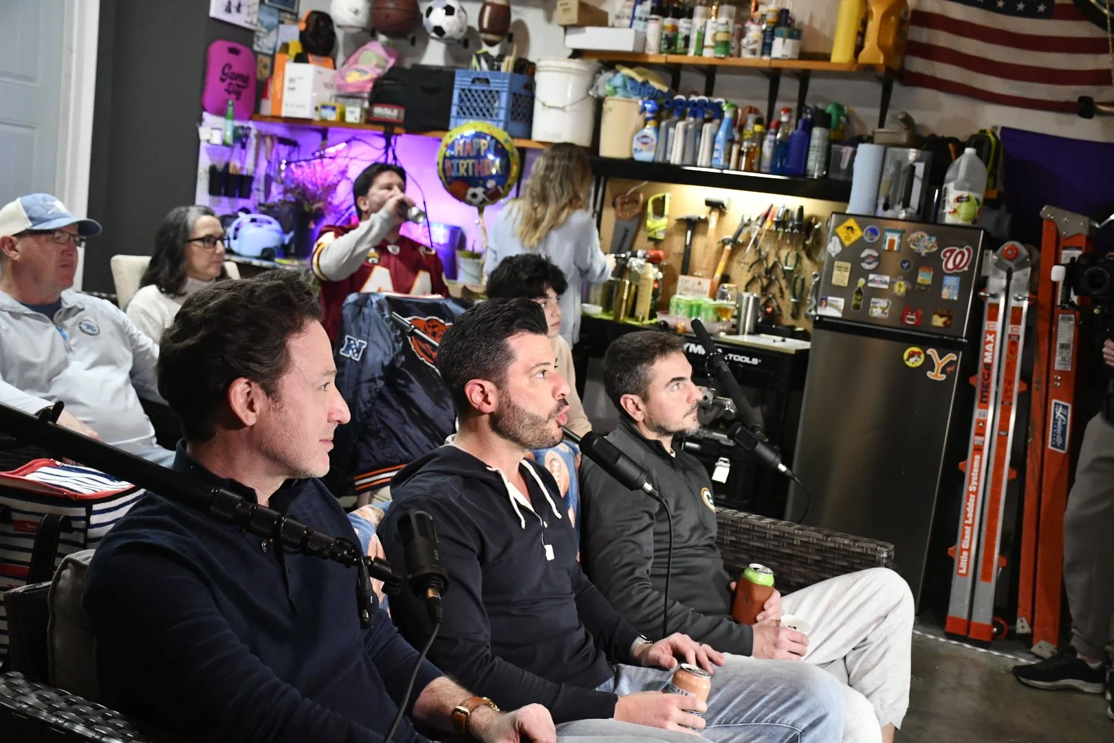 Group of men sitting in a garage, some holding drinks, attending a podcast or interview with microphones in front of them, with a woman in the background adjusting items on a workbench, surrounded by sports equipment, tools, and decorations.