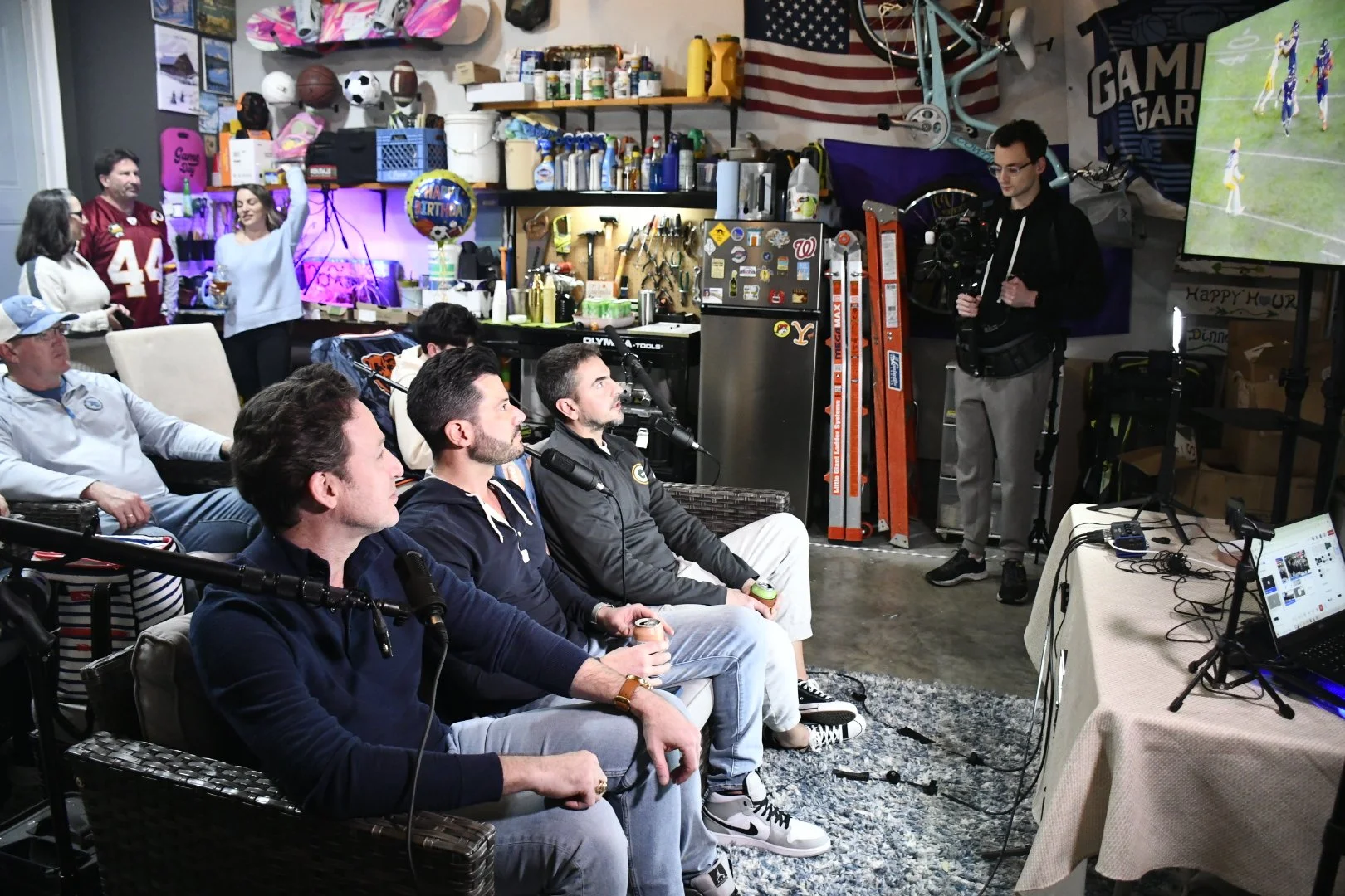 A group of people watching a sports game on a large TV in a garage-like space, with a camera operator filming them, shelves with tools and sports equipment, and festive decorations.